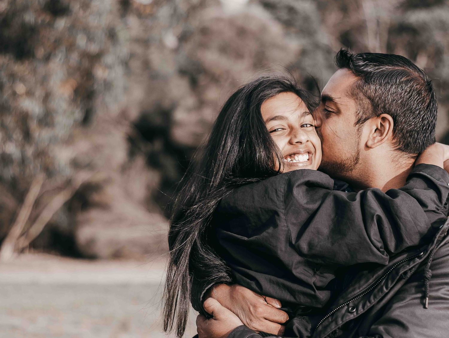 a young girl looking directly into the camera smiling whilst hugging a man who is kissing her on the cheek in front of trees