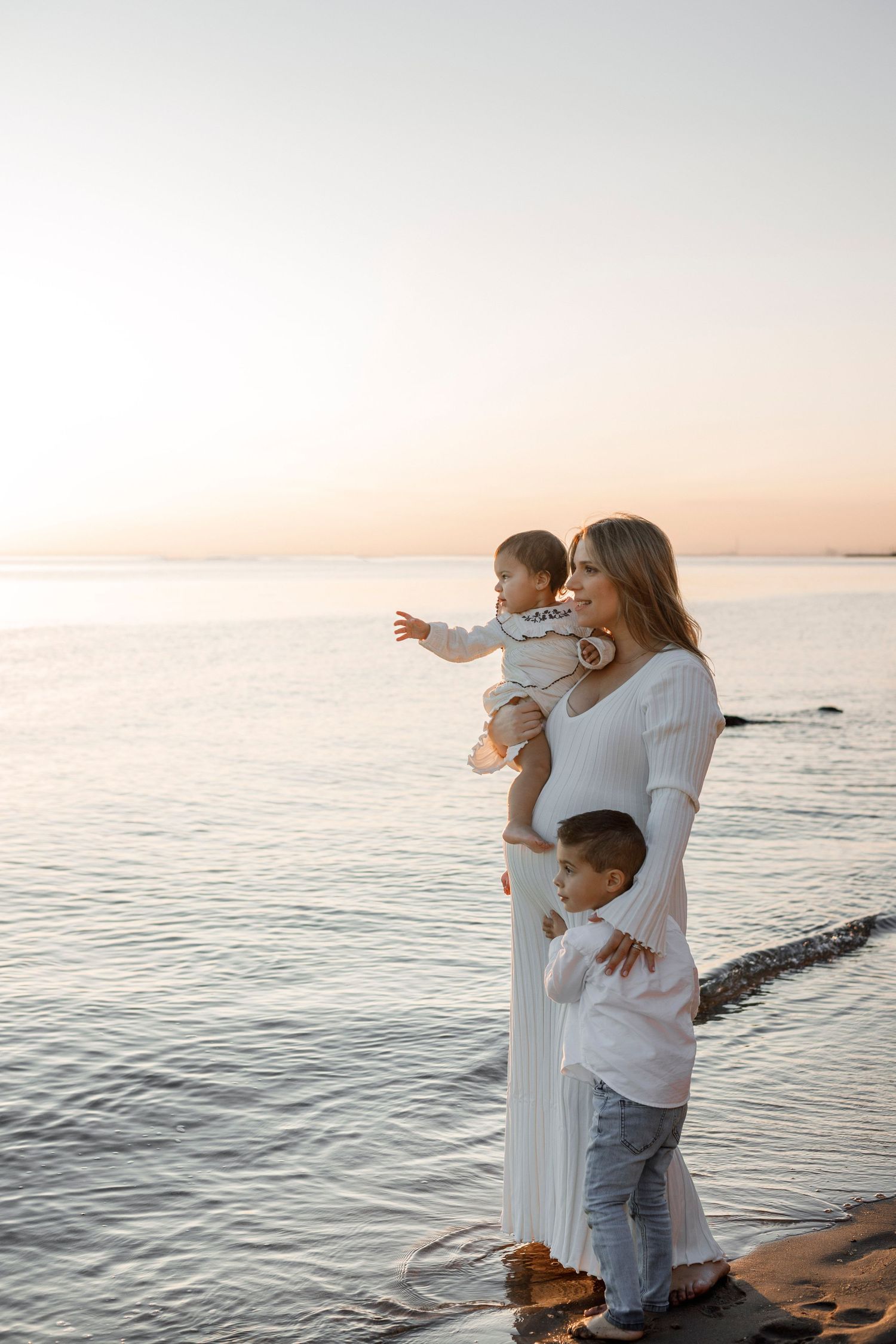 a pregnant woman wearing a white dress standing at the beach holding her two children  looking out into the distance with sunset light
