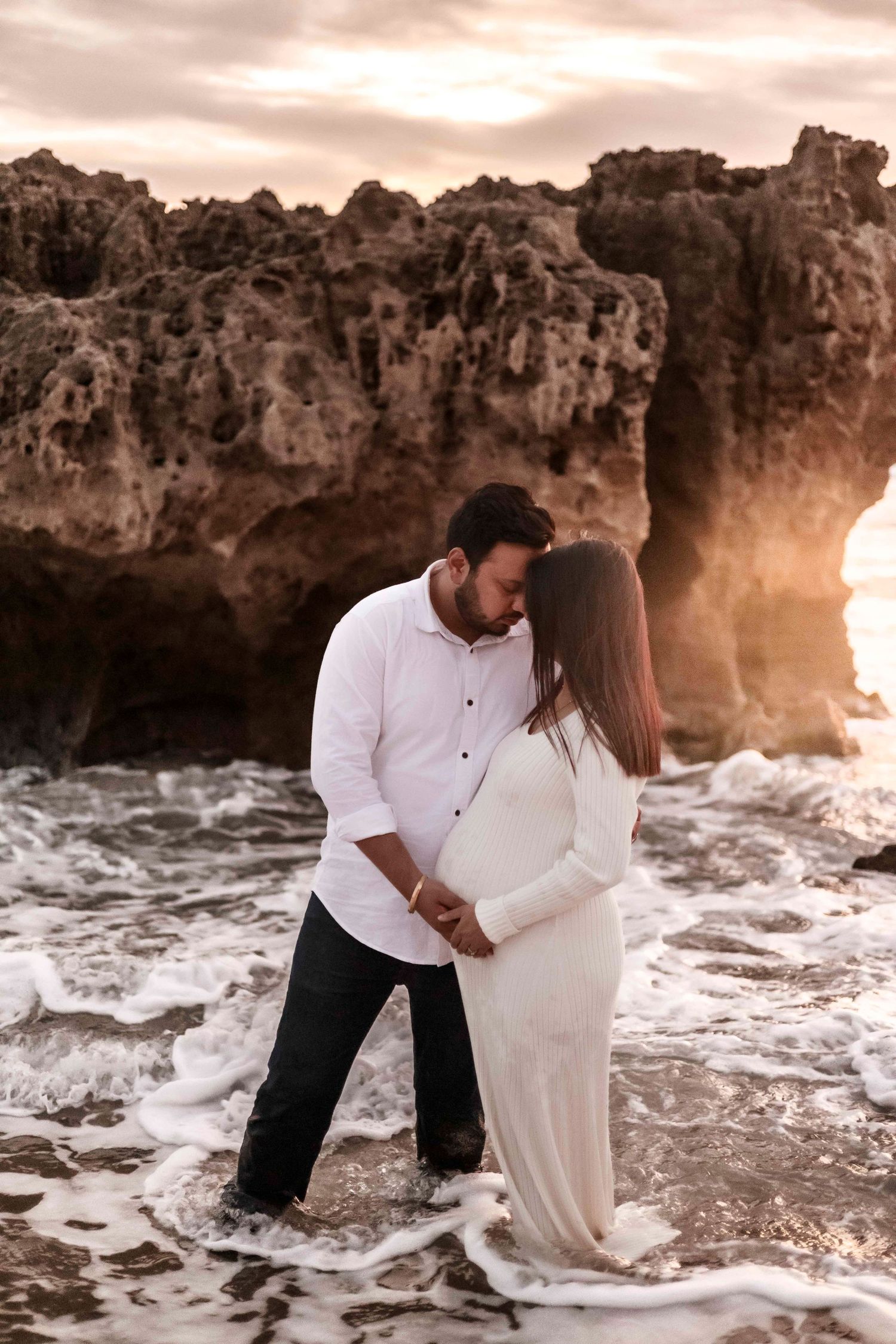 a man holding the belly of a pregnant woman wearing a white dress standing in the ocean with sunset light coming through rocks