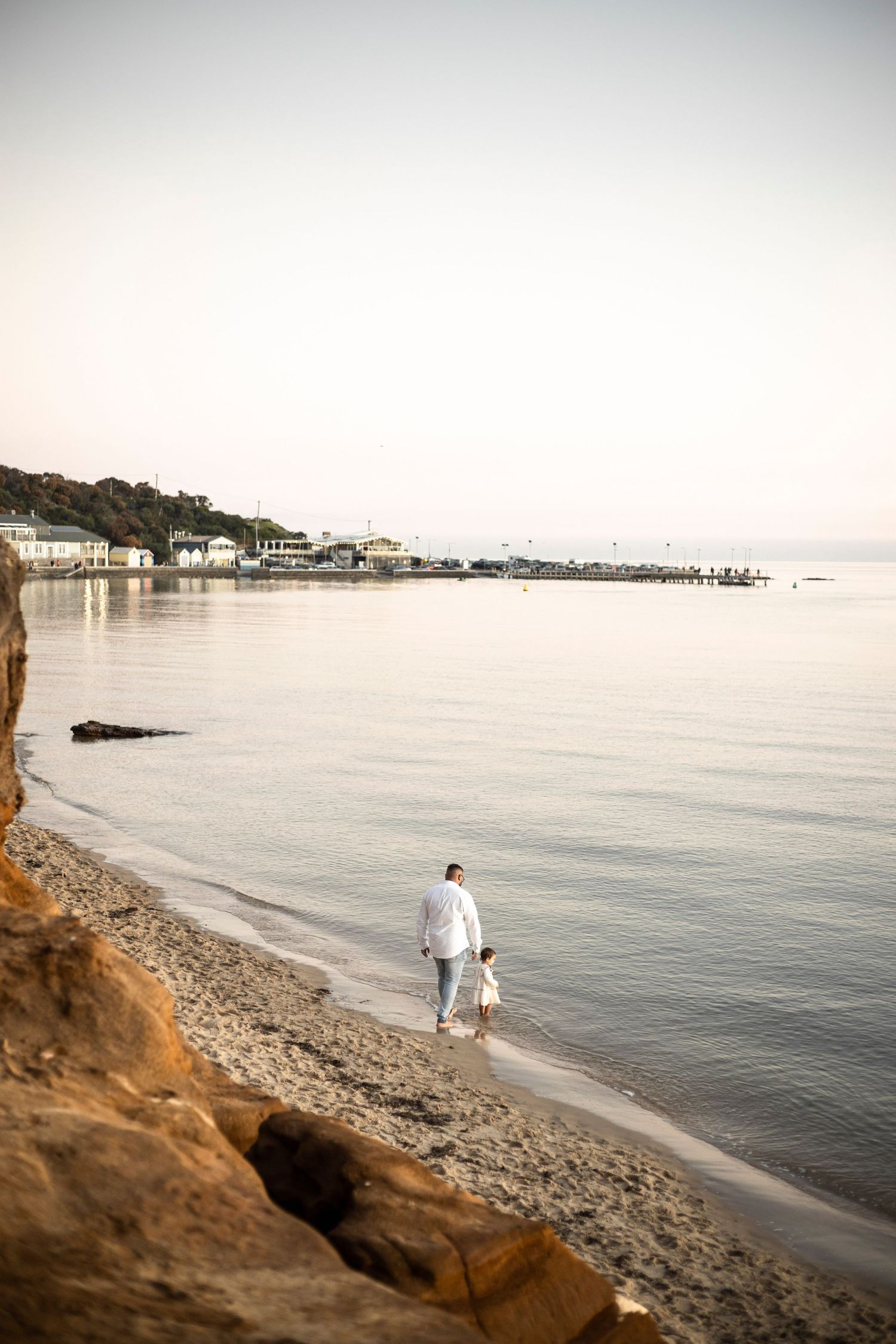 the back of a man and little girl standing on the sand at the beach looking into the ocean with the view of a jetty and rock cliff face