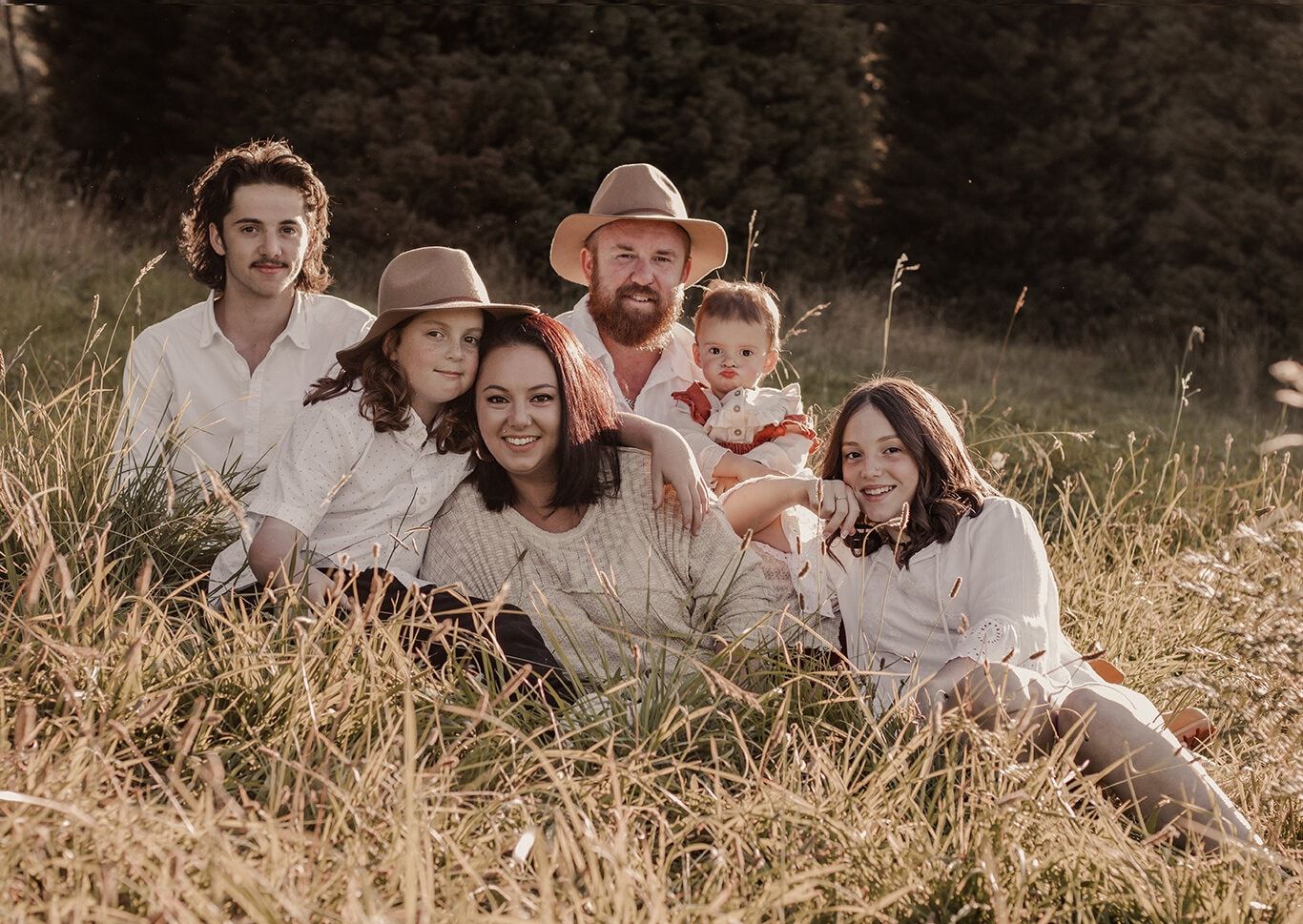 a man wearing a hat and a woman sitting on bright grass with four children looking directly at the camera wearing neutral coloured clothing