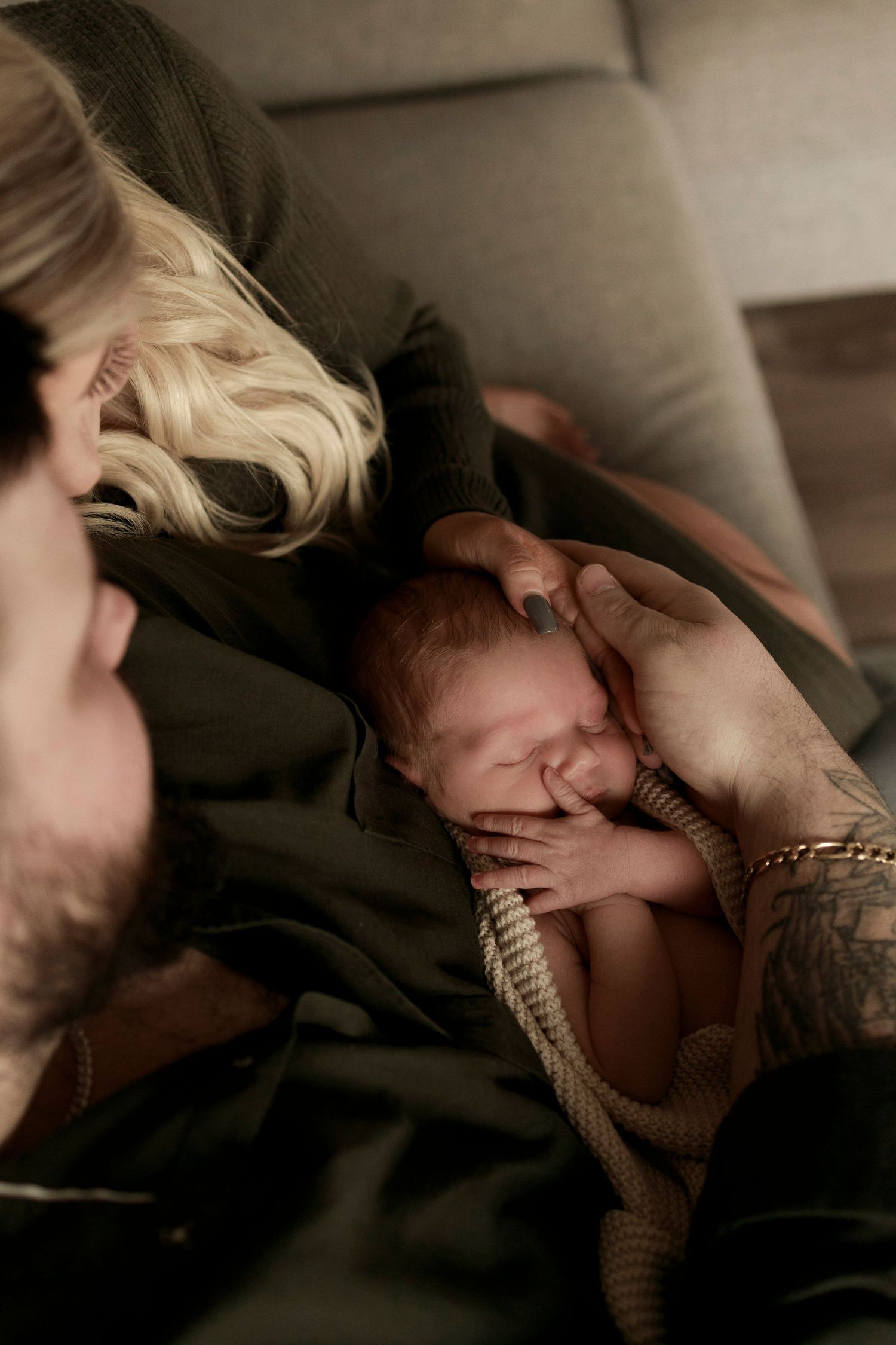 a newborn baby sleeping in a knitted beige blanket with hands showing and being held by mother and father as they look down at the baby