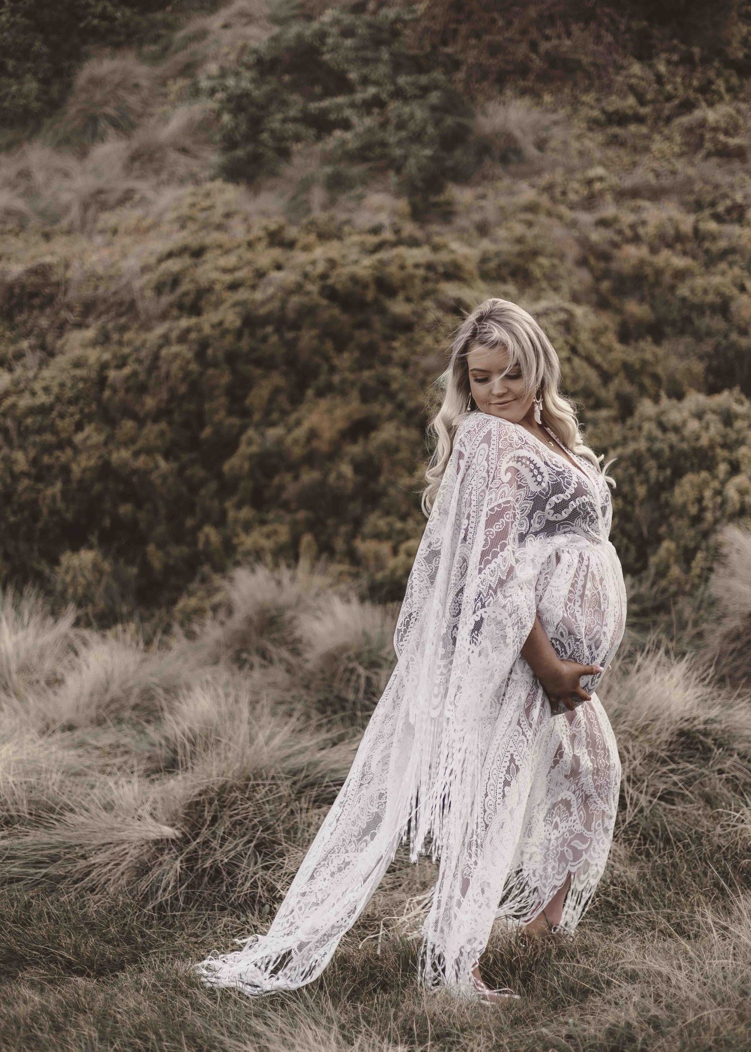 a pregnant woman wearing a white long lace dress holding her belly whilst standing amongst a grassy hill and looking down over her shoulder