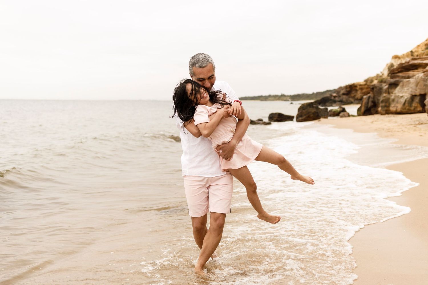 a man wearing pink shorts smiling carrying a young girl at a beach walking in the ocean next to rocks