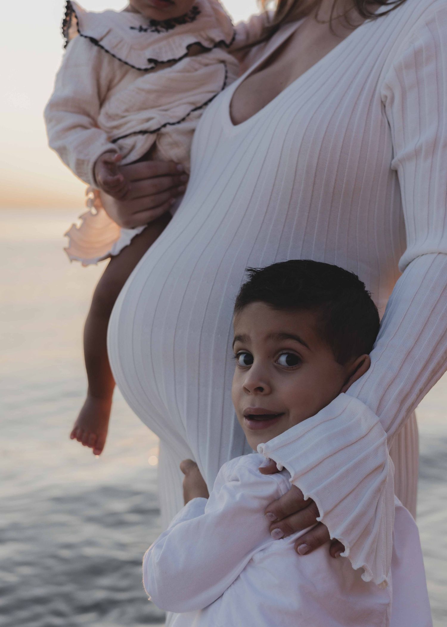 a pregnant woman standing at the beach with her child looking at the camera with her arm around him and holding another child on her hip