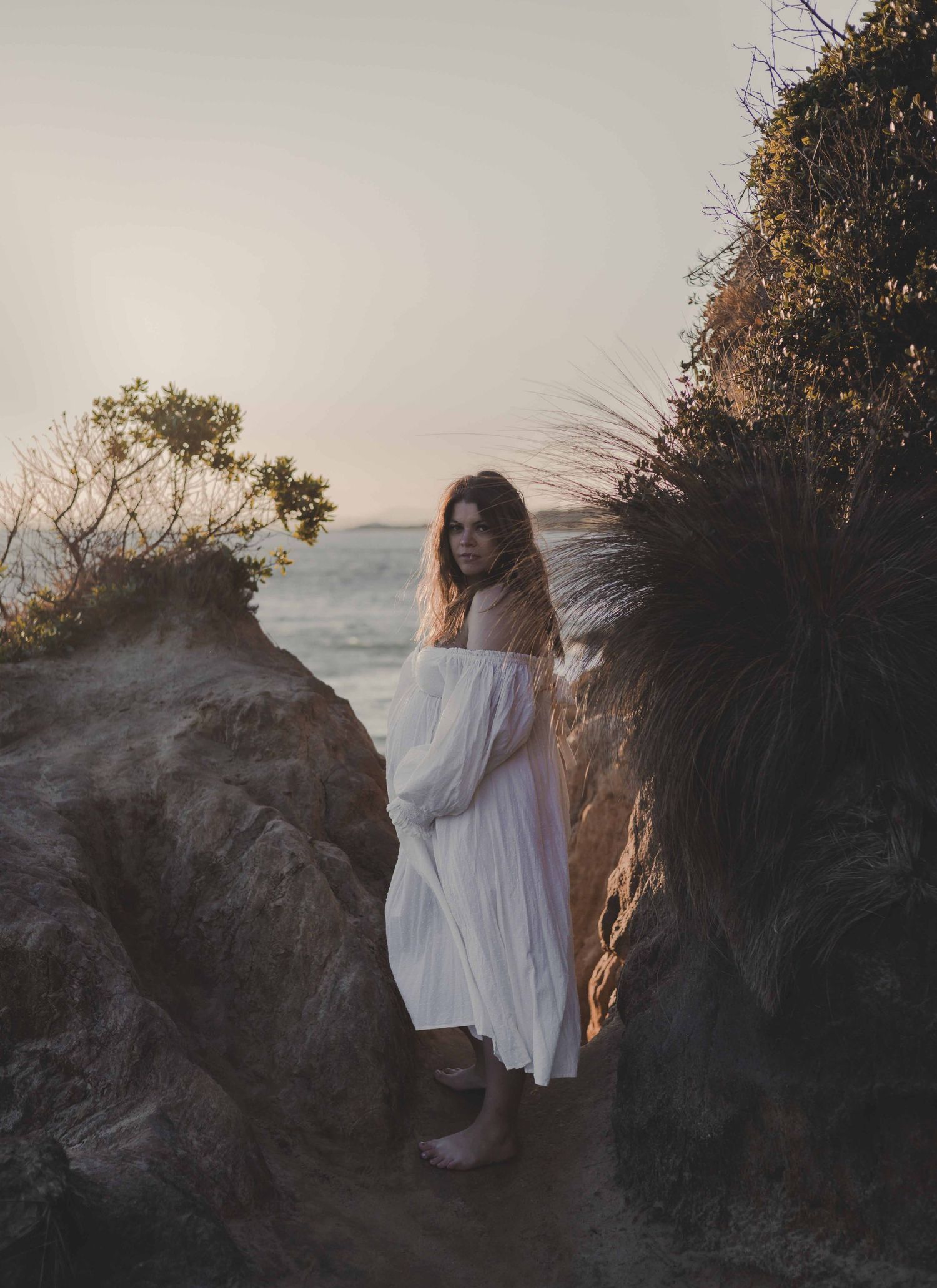 a pregnant woman standing amongst cliffs wearing a white dress whilst holding her belly and looking directly at camera