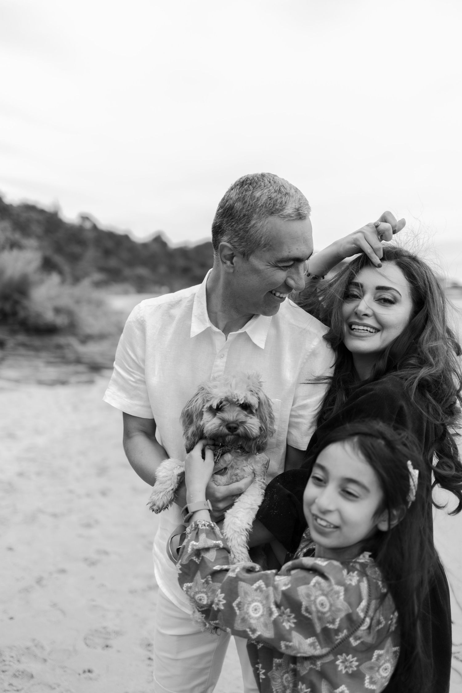 a woman smiling looking into the camera next to a man looking at her smiling whilst holding a dog and a smiling girl standing in front