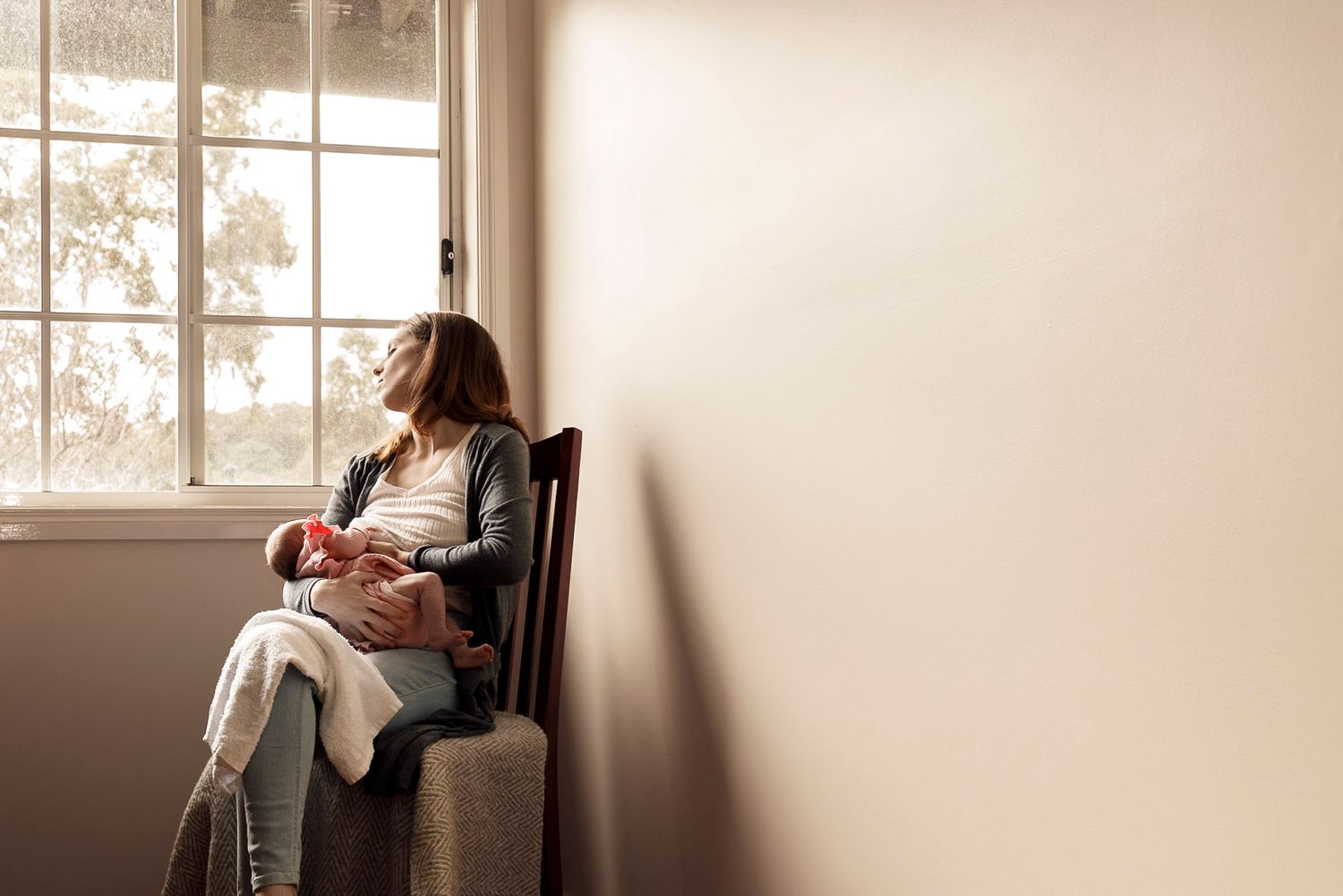a woman breastfeeding a baby sitting next to a window with eyes closed whilst facing towards the window