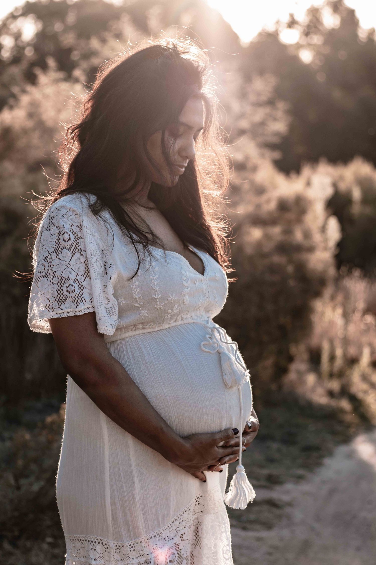 a pregnant woman with black hair standing outside in golden hour wearing a white dress whilst looking down and holding her belly