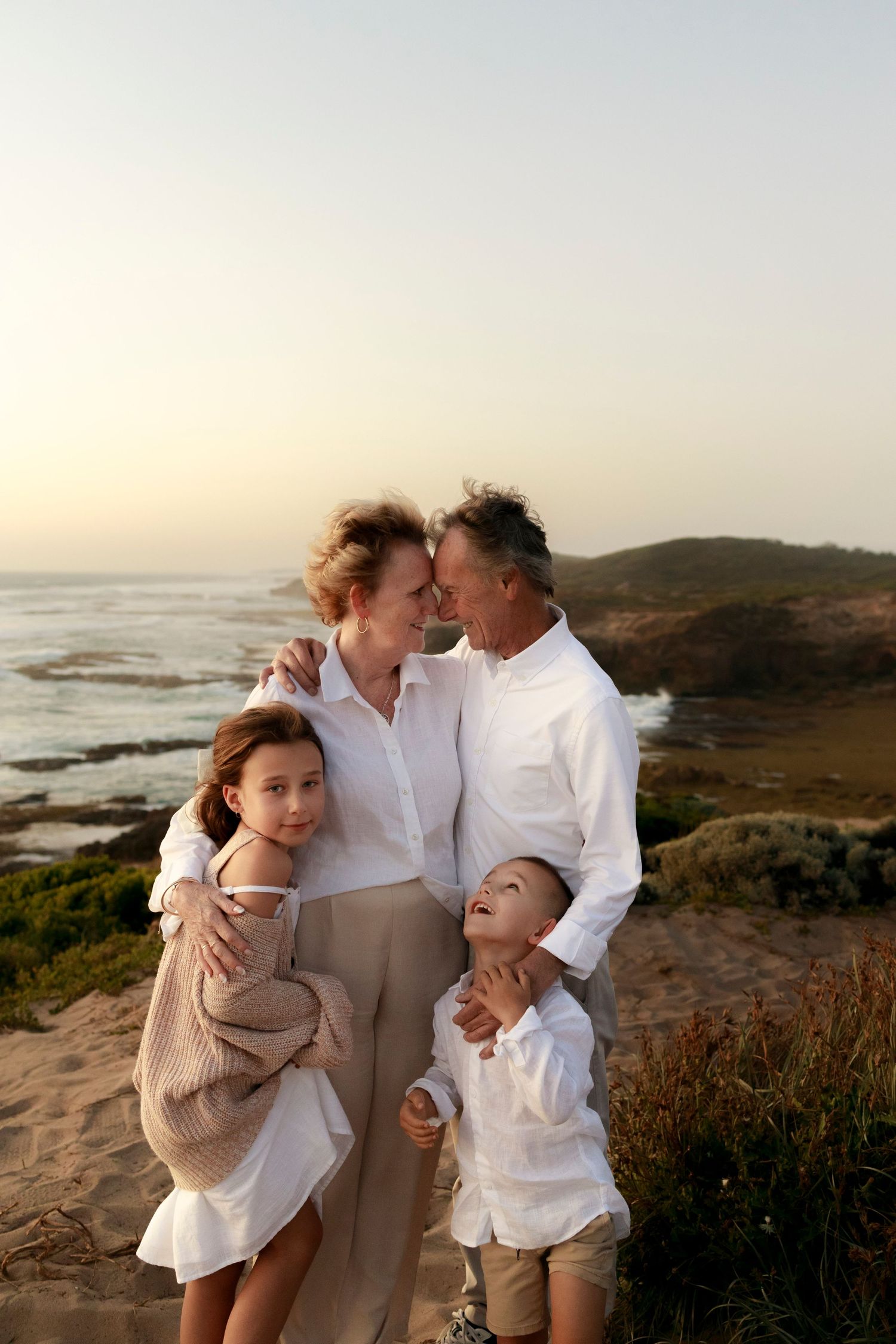 a man and woman wearing white standing with ocean behind them touching foreheads smiling with a girl and boy cuddling into them