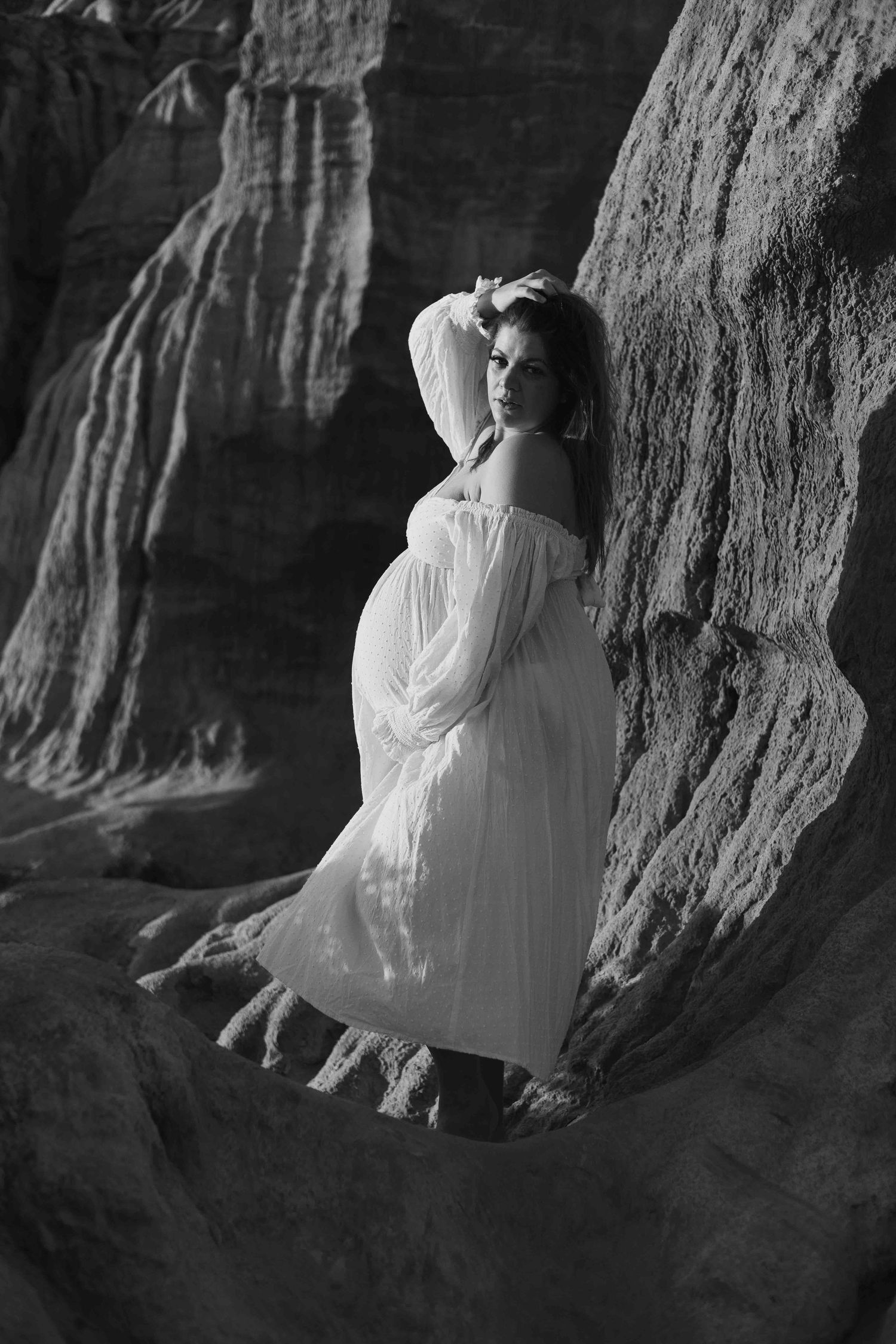 a pregnant woman standing in front of a rock cliff face holding her hair up and looking directly into the camera wearing a white dress