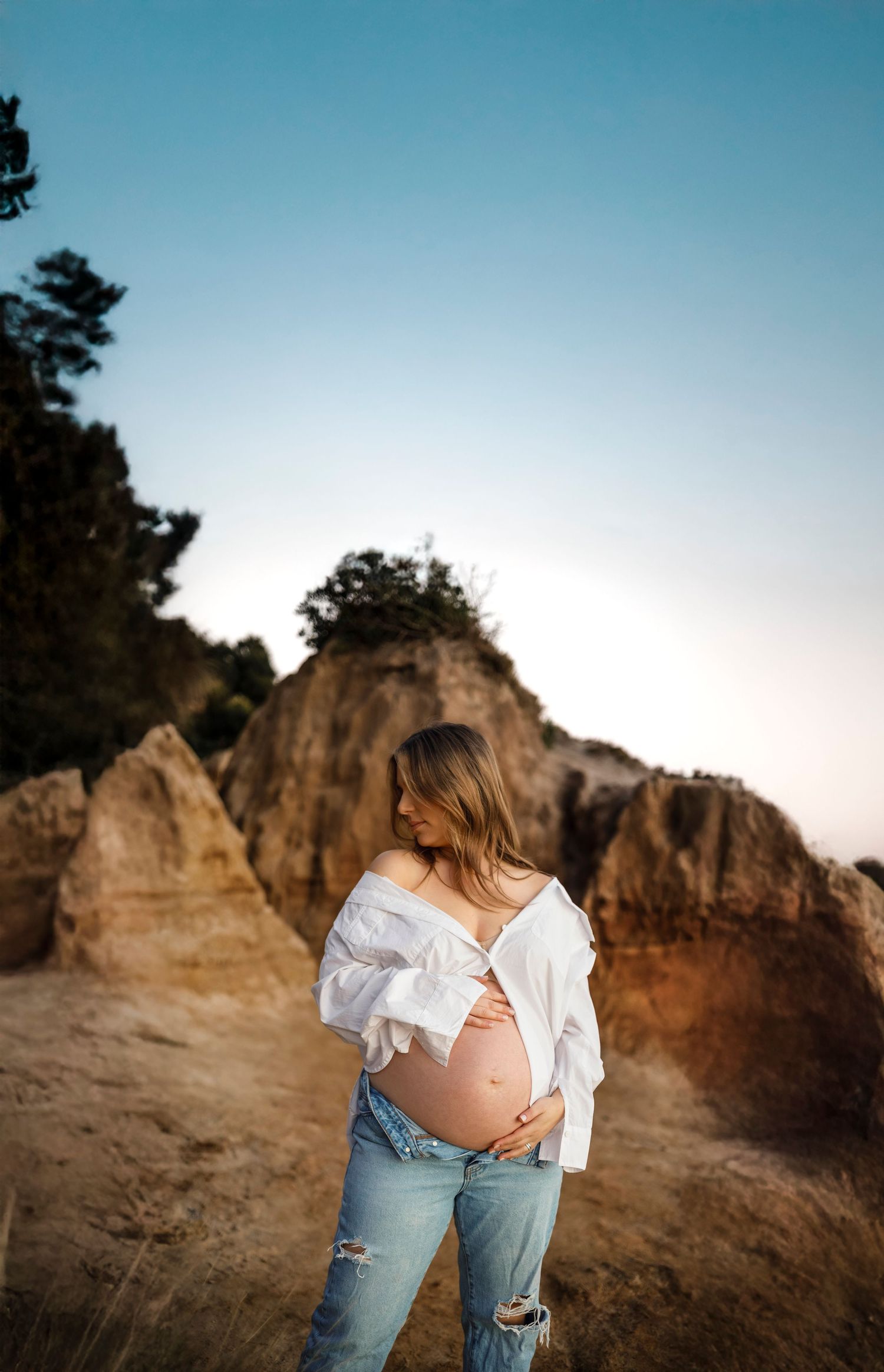a pregnant woman standing in front of a cliff wearing a white shirt and blue jeans whilst looking over her right shoulder with eyes closed