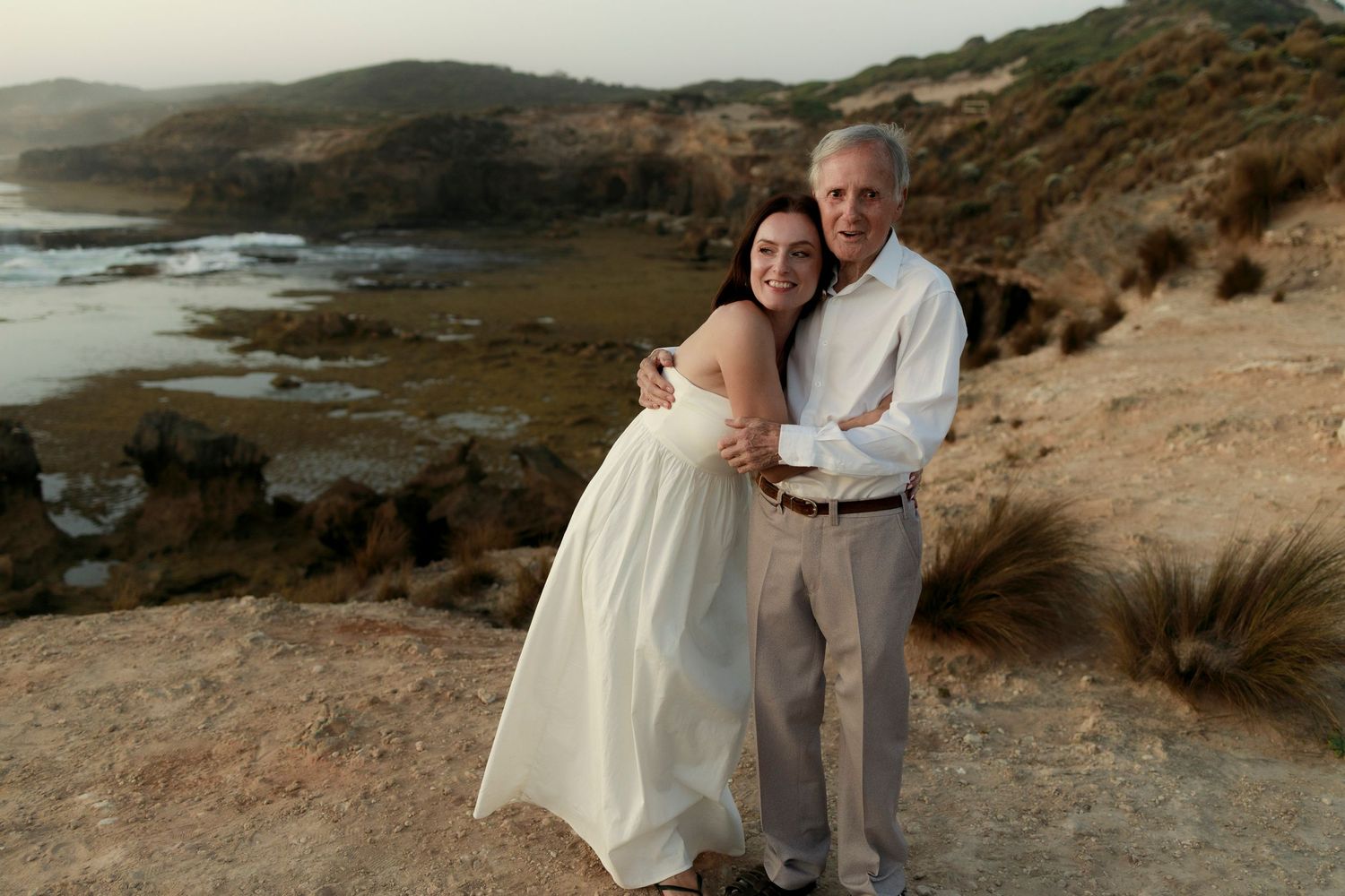 an elderly man wearing a white shirt standing on a cliffside looking directly into camera with a woman hugging into him and smiling