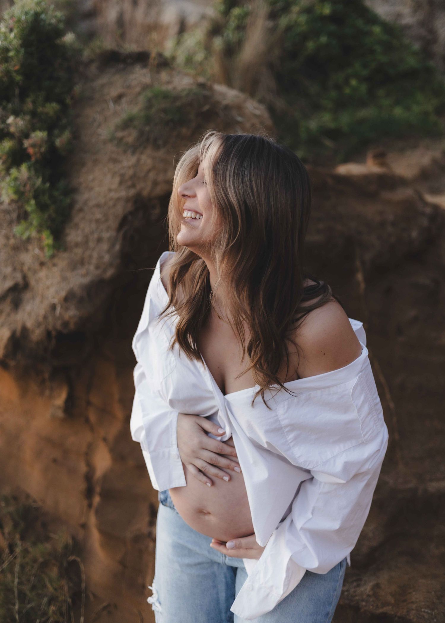 a blonde pregnant woman holding her belly whilst leaning over and smiling wearing white shirt and blue jeans