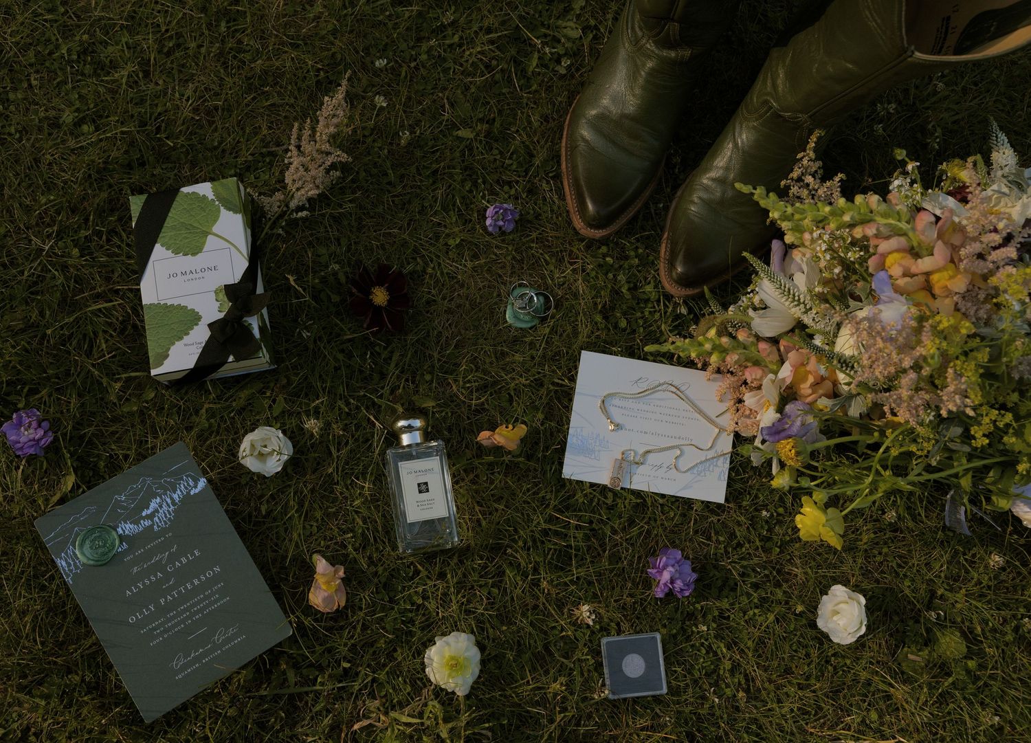 Flat lay of wedding stationery, Jo Malone perfume, wildflowers, boots, and jewelry on green grass.