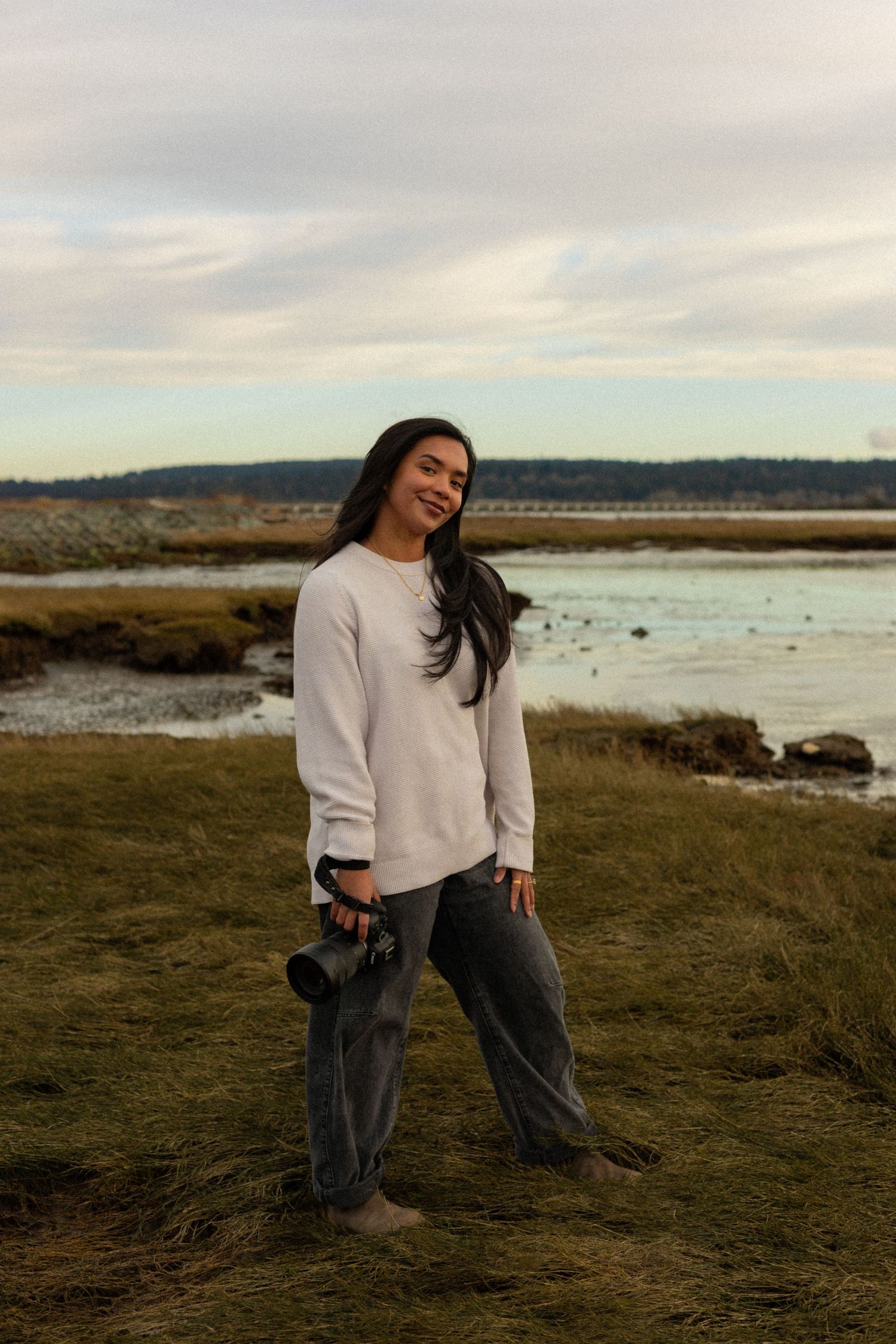 Smiling woman in a cream sweater and gray pants holds a camera while standing on a grassy shoreline at dusk beside calm tidal water.