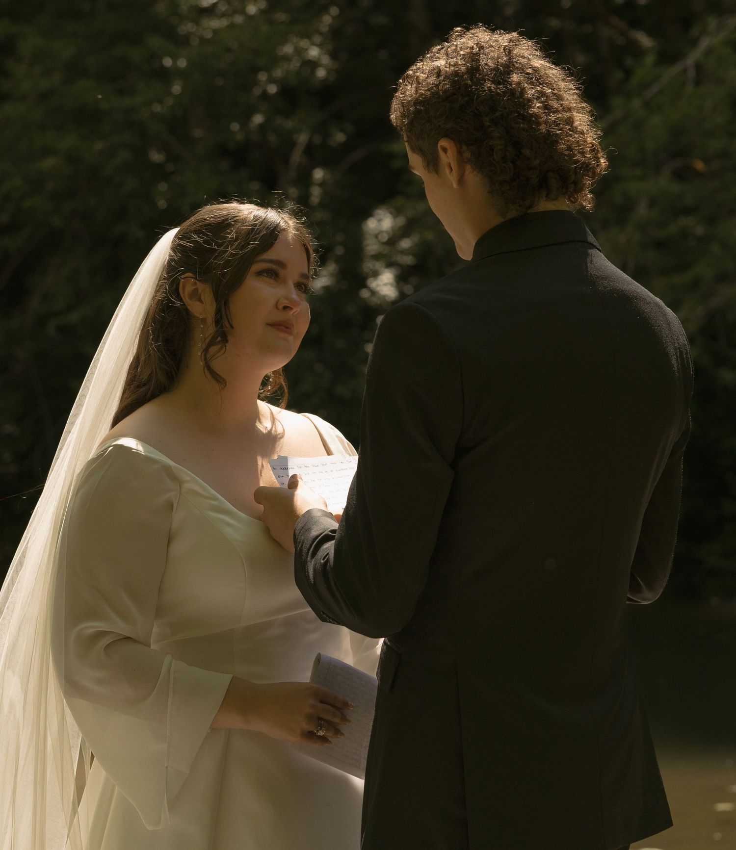 Bride in white gown and veil exchanges vows with groom in black suit during outdoor wedding ceremony.
