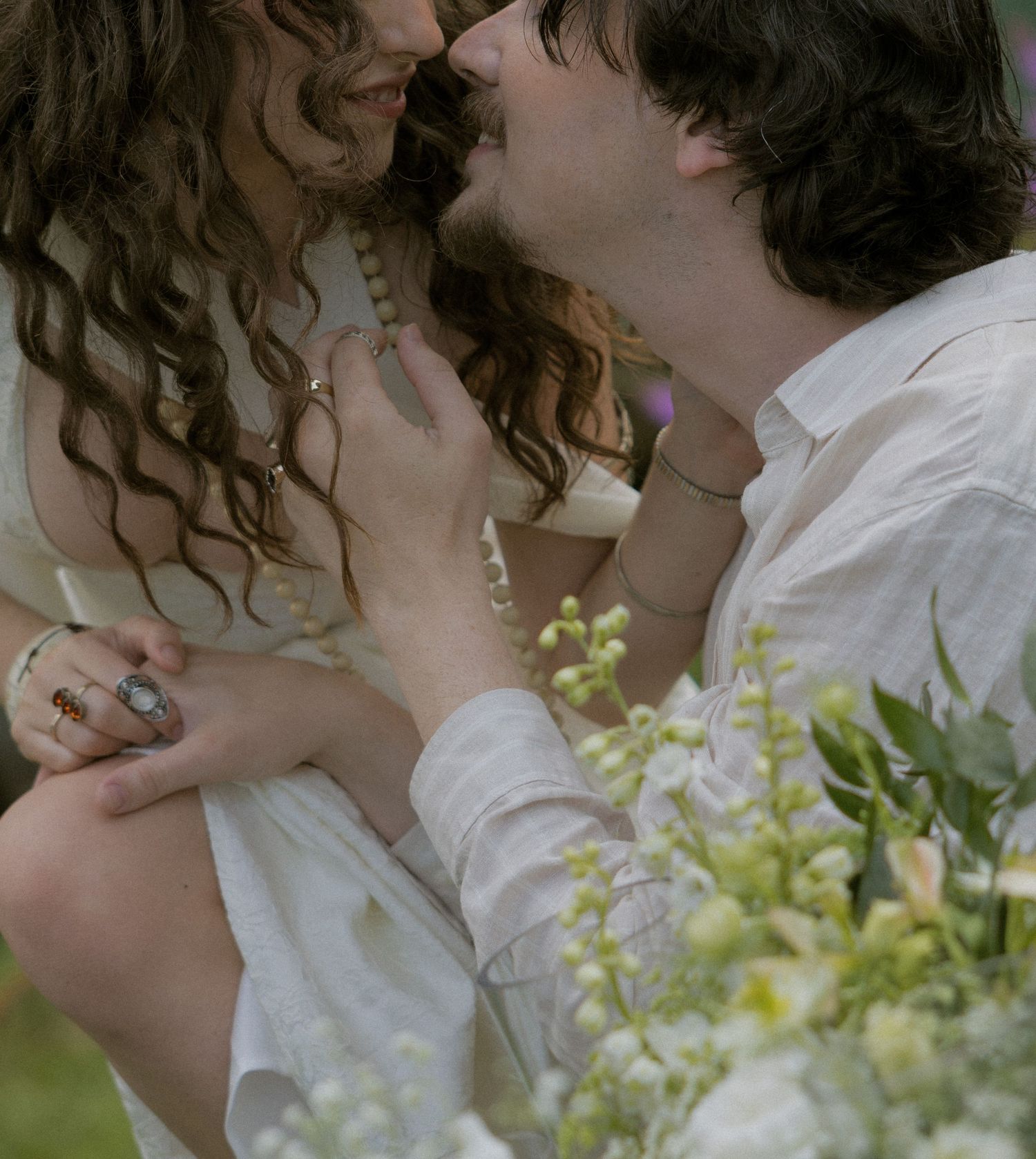 A romantic couple in white bohemian attire sharing a tender kiss surrounded by delicate white flowers outdoors.