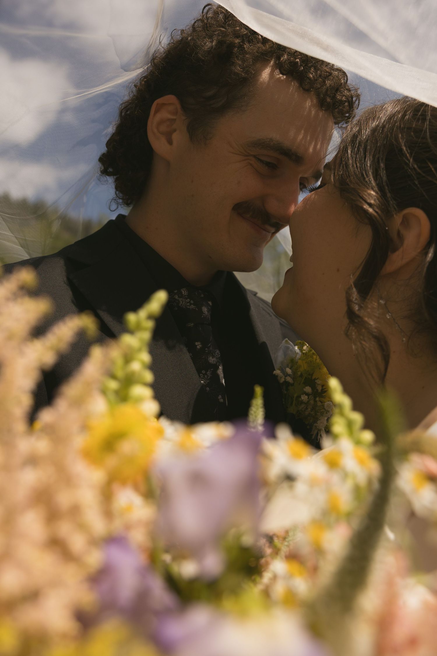 A bride and groom share a tender moment under a veil, surrounded by colorful wildflowers at their outdoor wedding in Squamish, BC