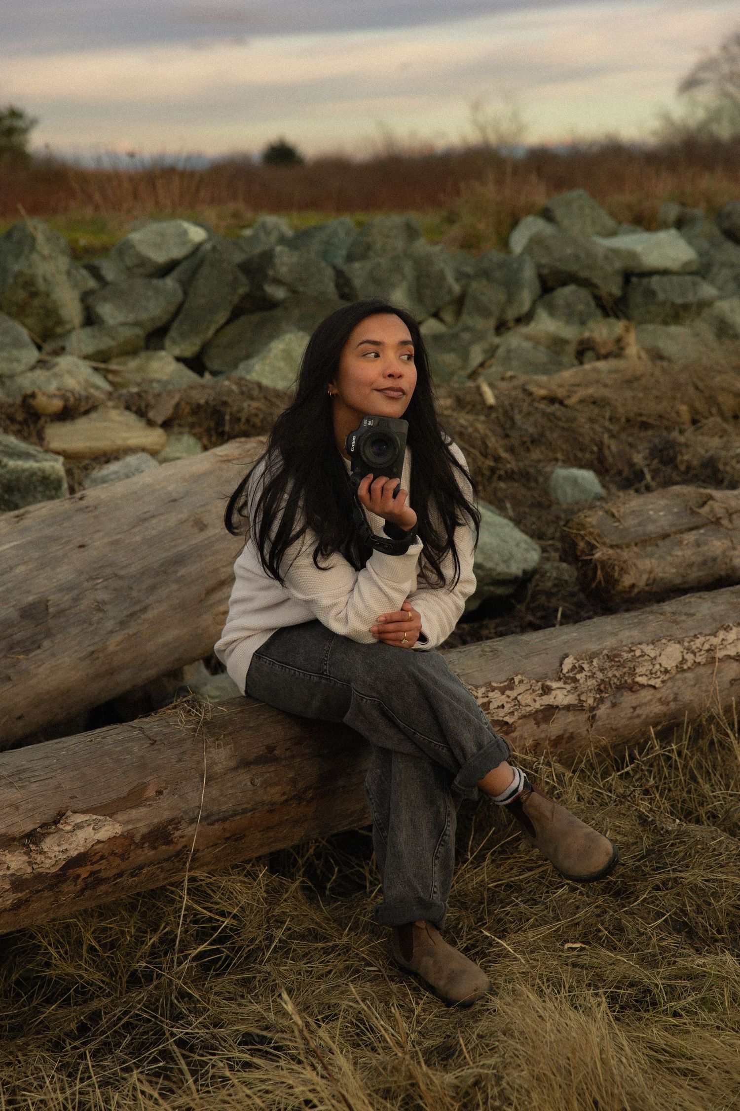 Young woman sitting on driftwood logs holding a camera, surrounded by rocks and dry grass at dusk.