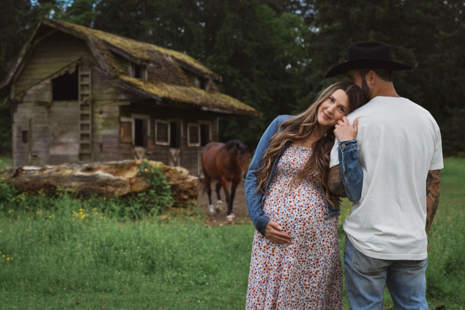 A couple in casual attire poses together in a grassy outdoor setting during a maternity photo session.