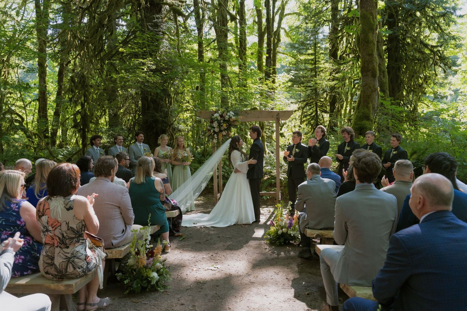 Outdoor forest wedding ceremony with guests seated among tall trees as couple exchanges vows at a floral arch.