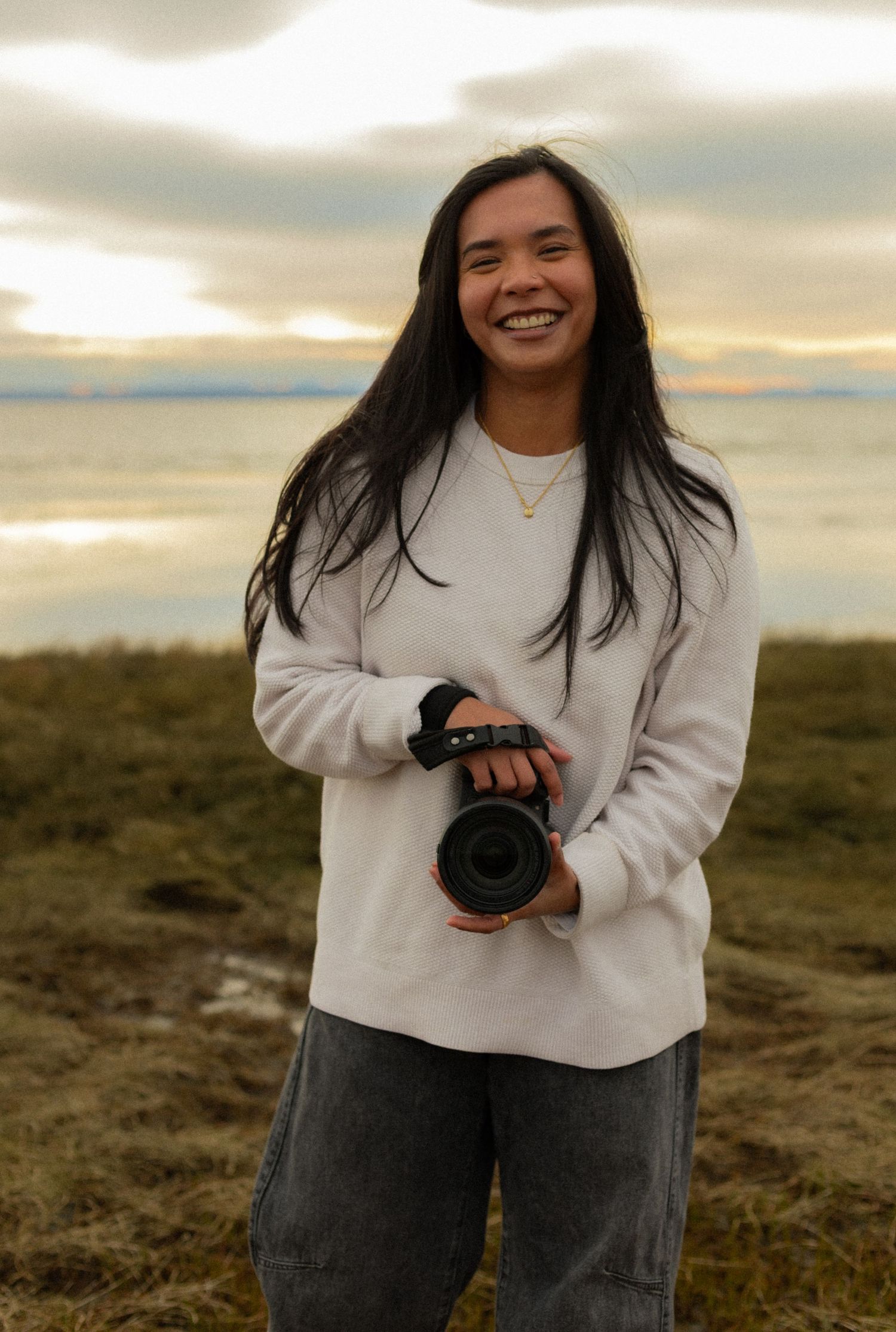 Photographer in white sweater and jeans holds camera while laughing during golden hour photoshoot in grassy field.