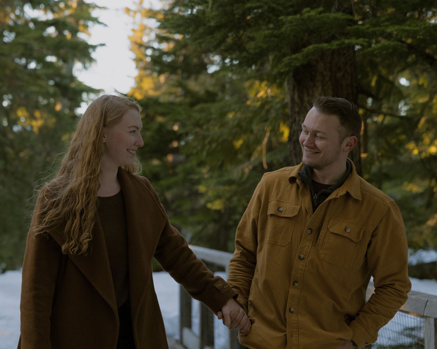 Couple holding hands and smiling at each other outdoors in a snowy winter forest setting.
