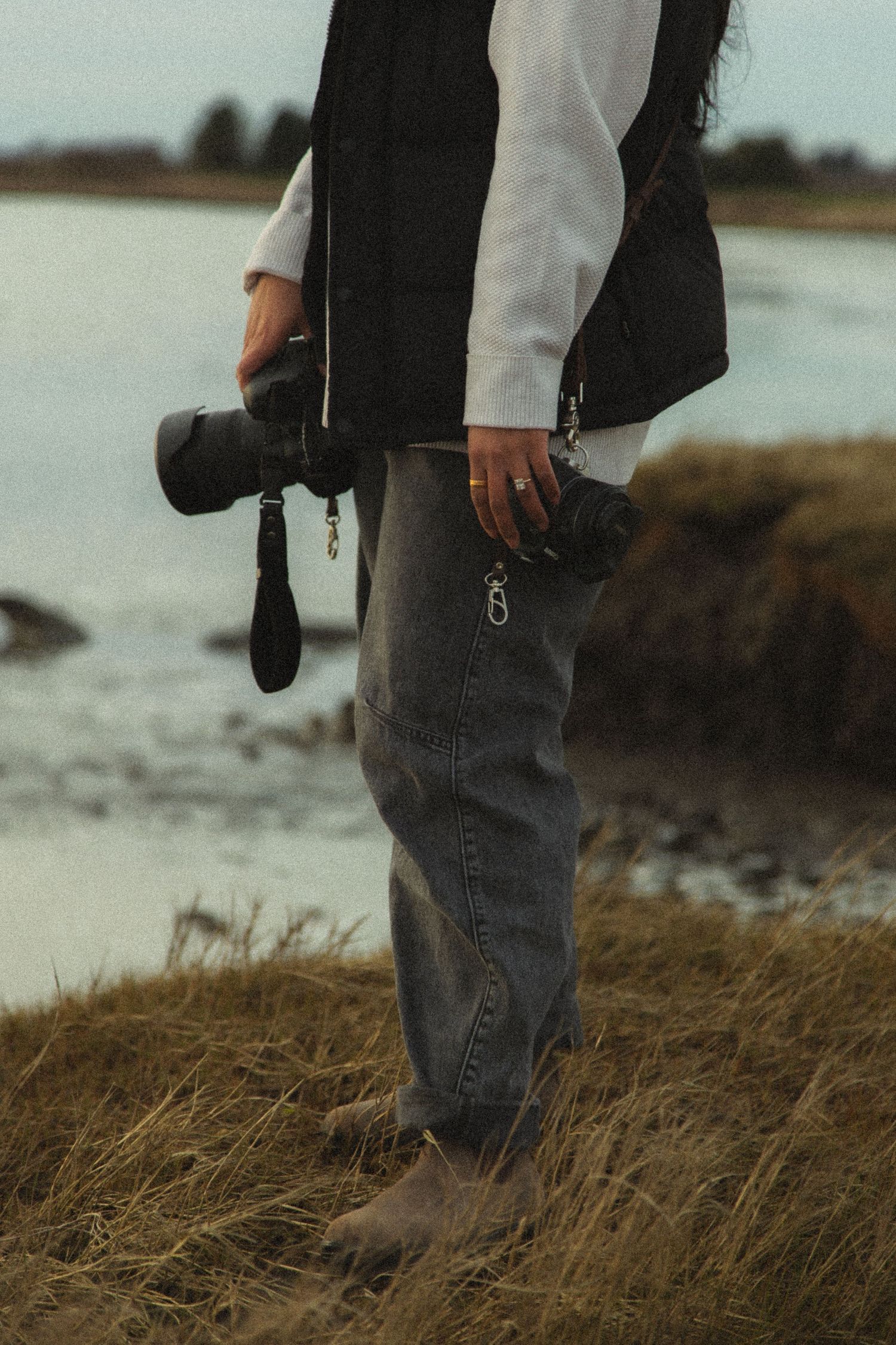 Barefoot photographer standing in coastal grass holding a DSLR camera, wearing jeans and black vest.