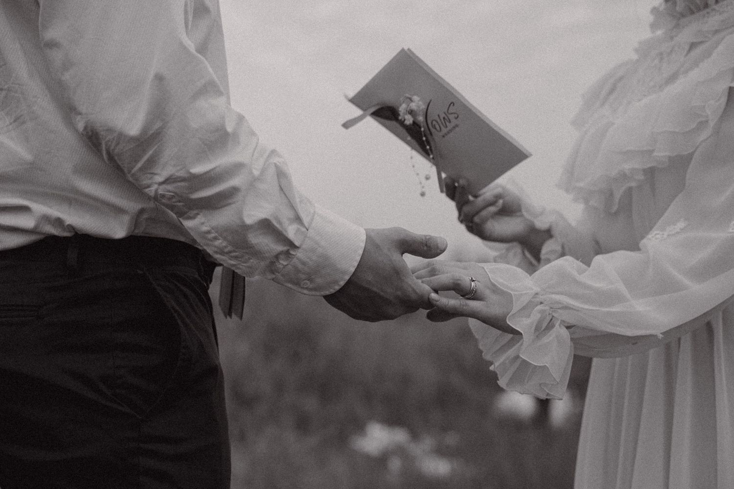 Black and white photo of a couple exchanging rings during an outdoor wedding ceremony.