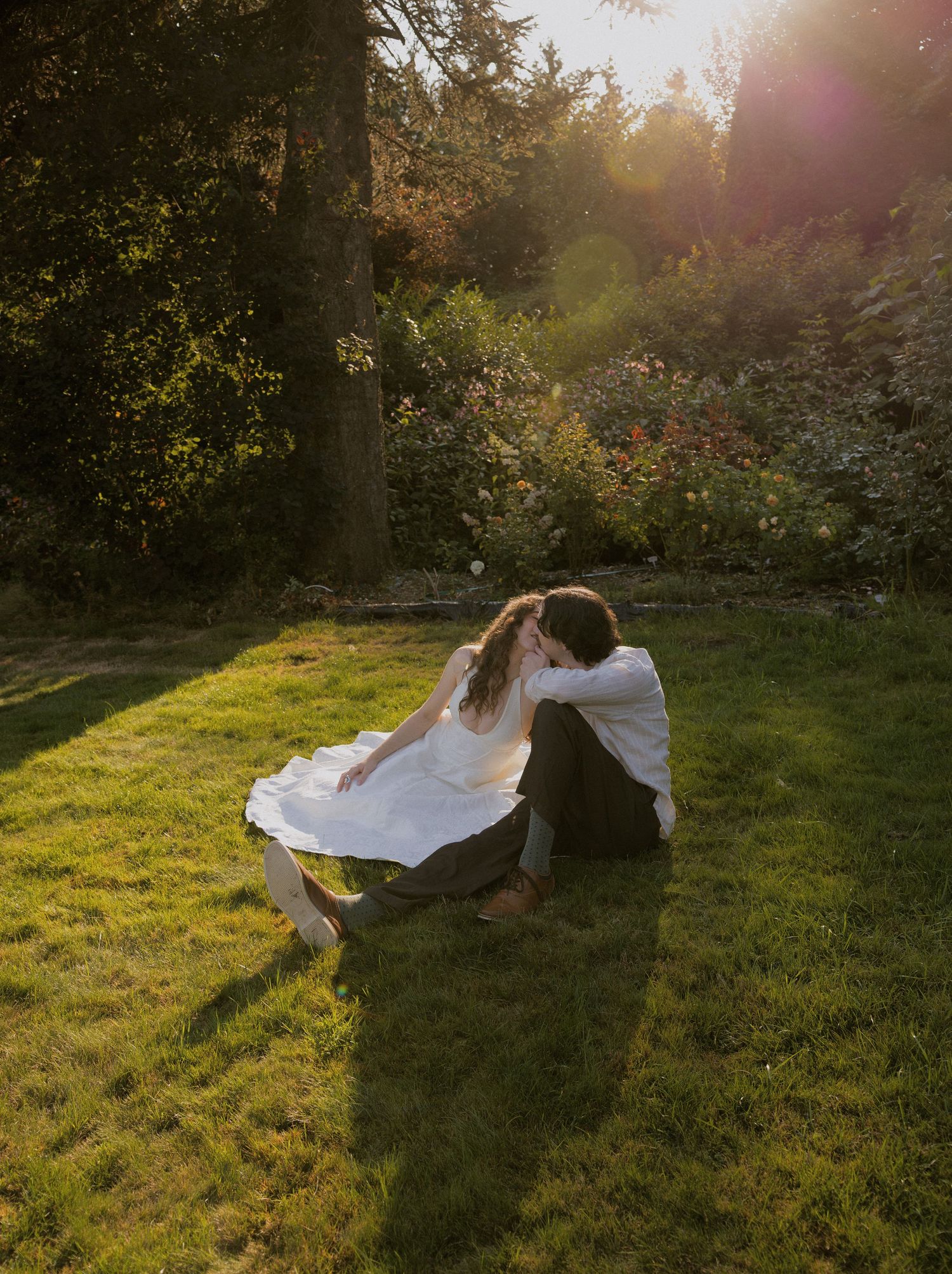 Couple sharing a romantic embrace on green grass at golden hour, surrounded by lush garden trees and warm sunlight.