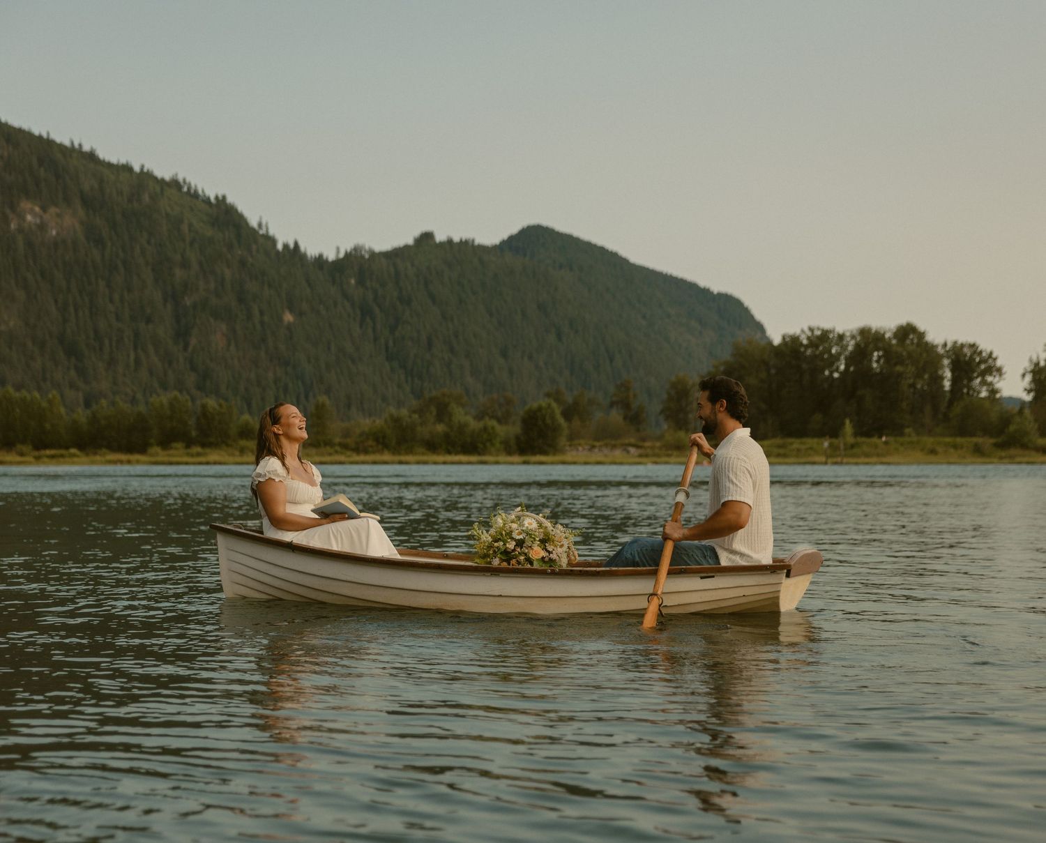 Bride and groom rowing a wooden canoe on a calm mountain lake, with lush green trees and hills in the background in Pitt Meadows, BC