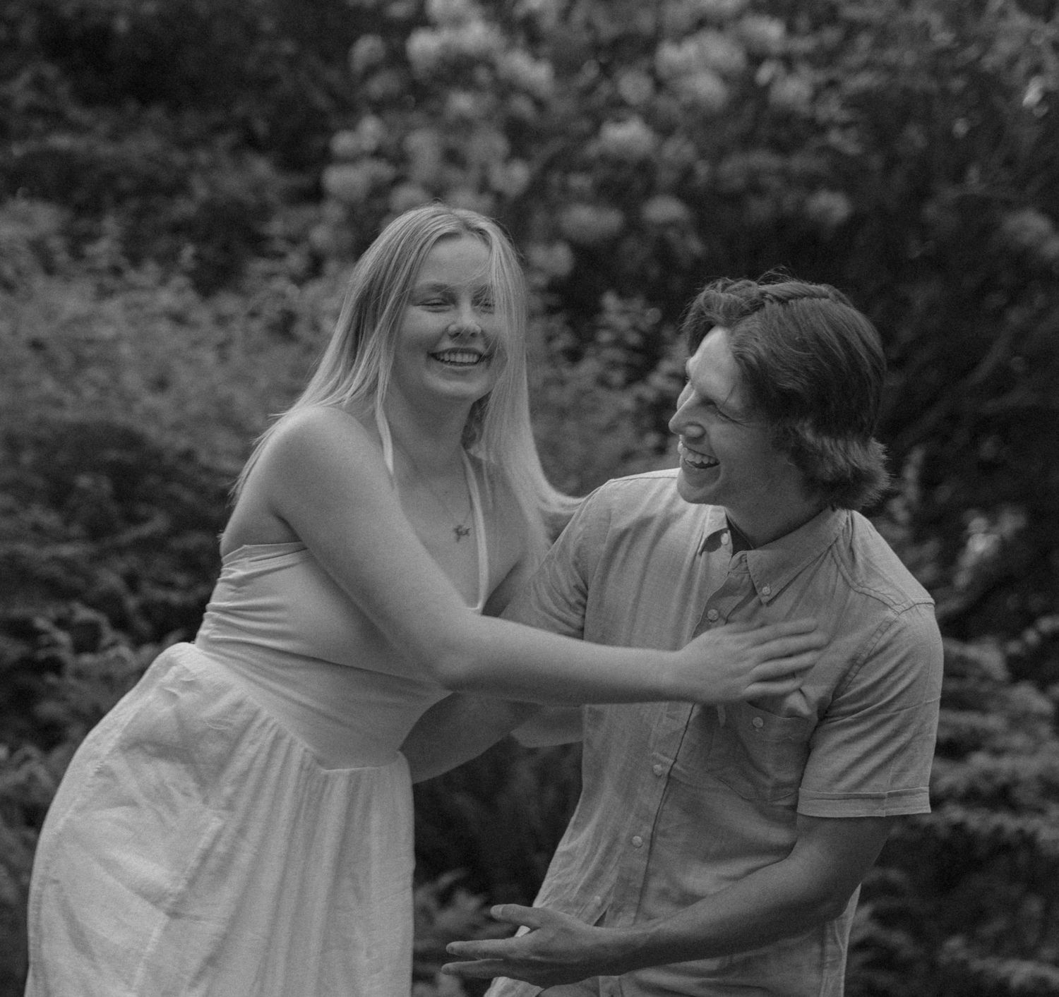 Black and white photo of a laughing couple embracing outdoors with lush garden greenery in the background.