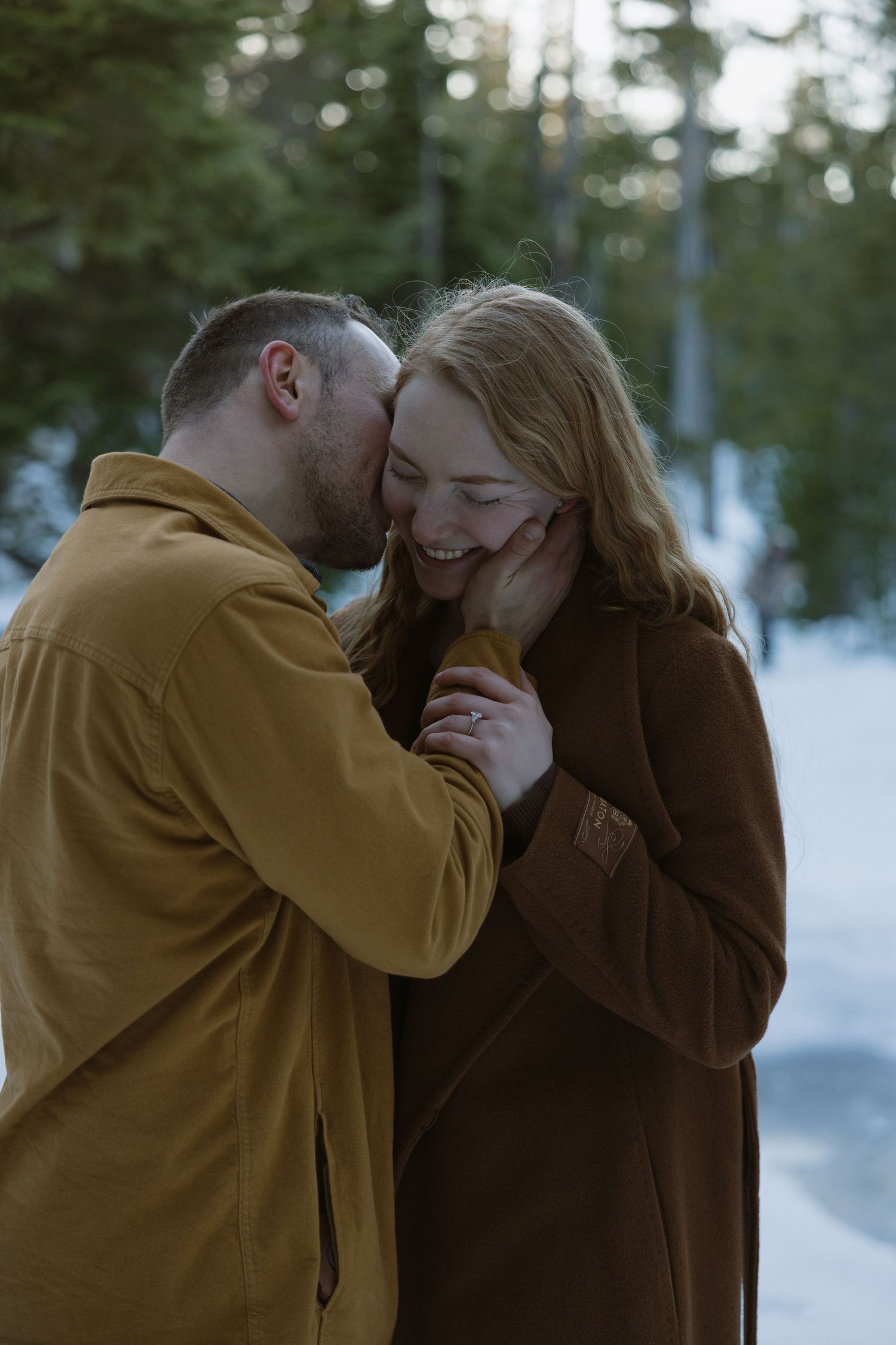 A couple shares a tender moment in a snowy forest, the man kissing her cheek as she laughs, both wearing warm brown coats.