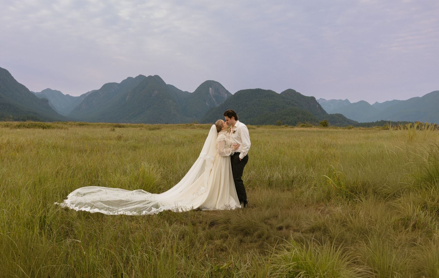 Bride and groom share a romantic kiss in a vast green meadow with dramatic karst mountains in the background.