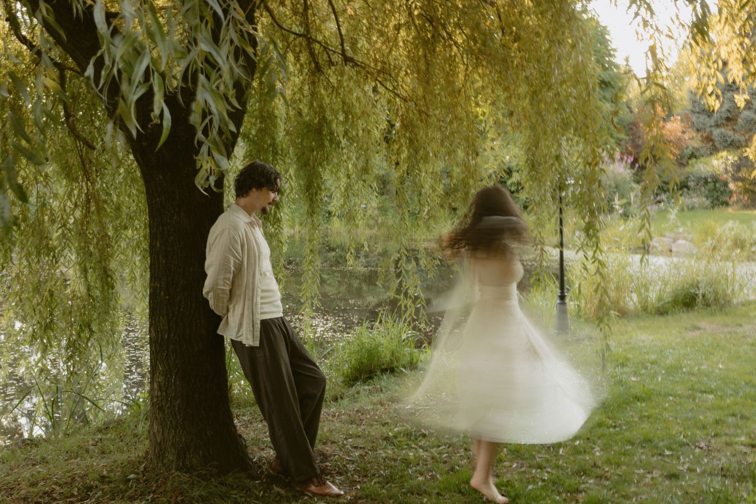 A couple shares a romantic moment under a weeping willow tree by a serene pond, the woman twirling in a white dress.