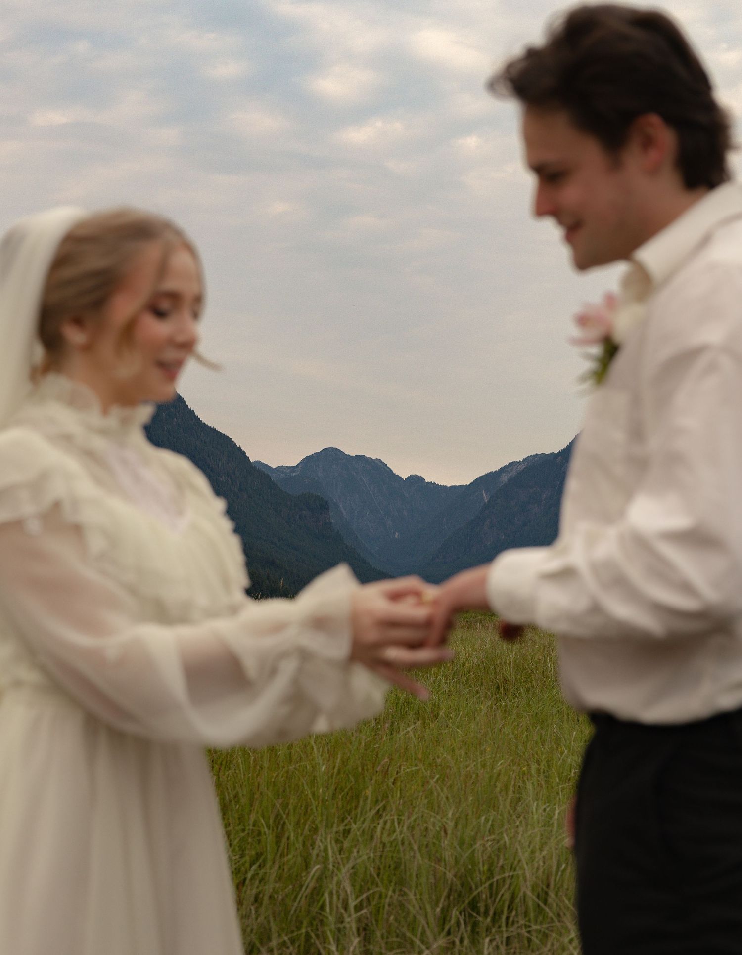 A bride and groom hold hands in a meadow with mountain backdrop, wearing white wedding dress with veil and boutonniere.