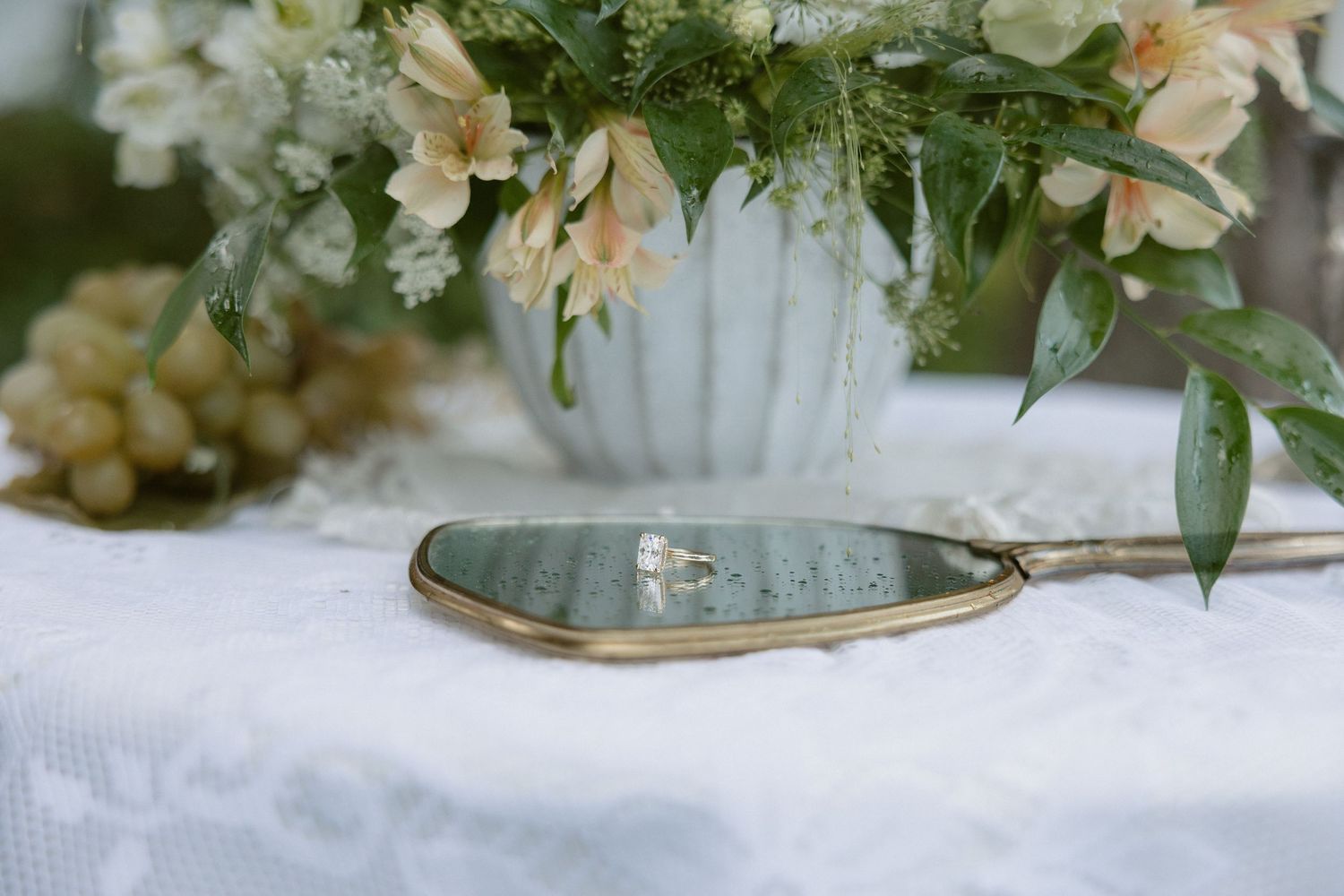 Gold cake server with wedding rings on marble dish beside floral arrangement and grapes on lace tablecloth.