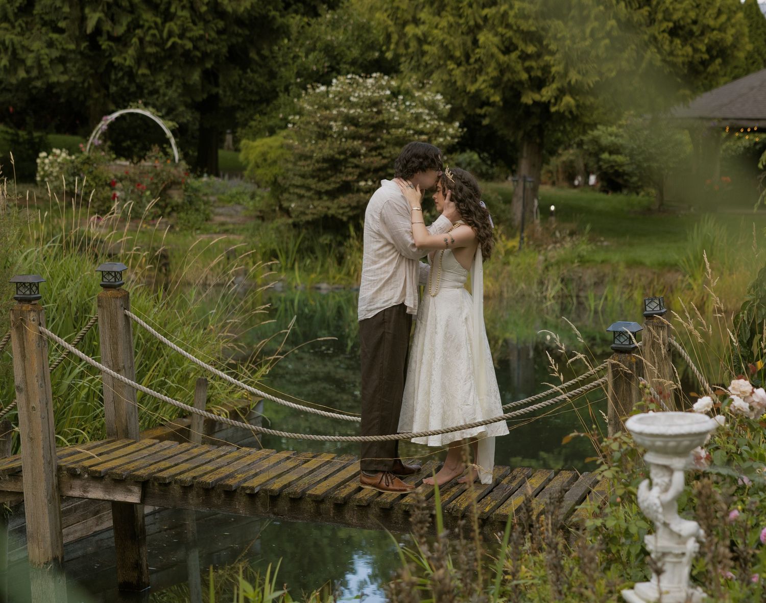 Couple embracing on a wooden bridge over a pond in a lush green garden setting.