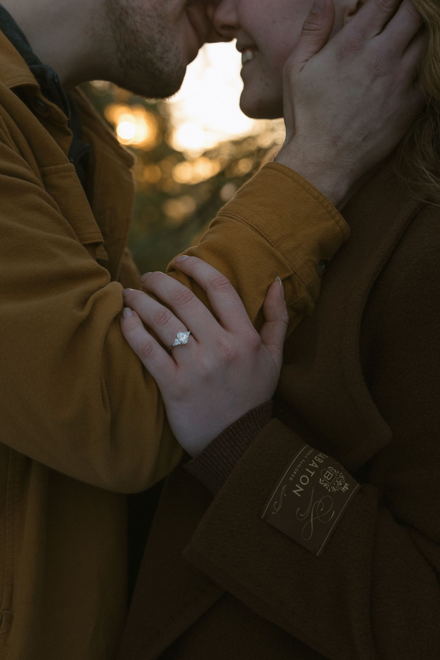 Couple in matching brown jackets sharing a romantic moment, woman wearing a diamond engagement ring.