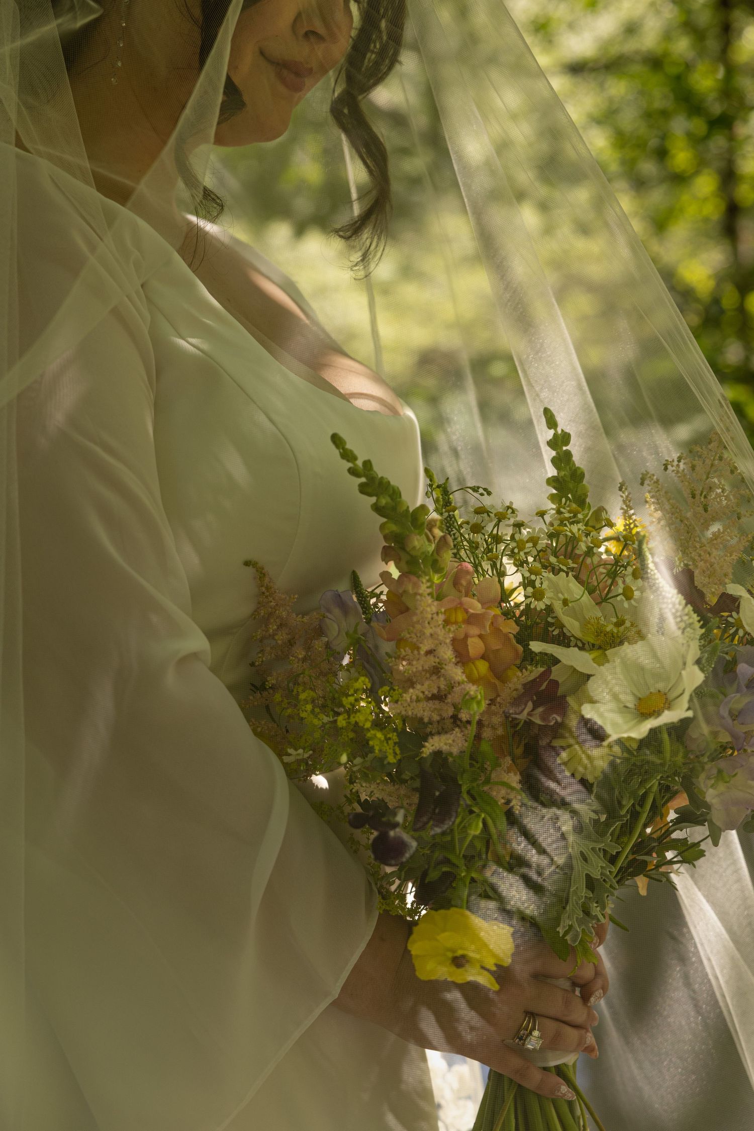 Bride in white gown and veil holding a wildflower bouquet with yellow and purple blooms in a sunlit garden setting in Squamish, BC