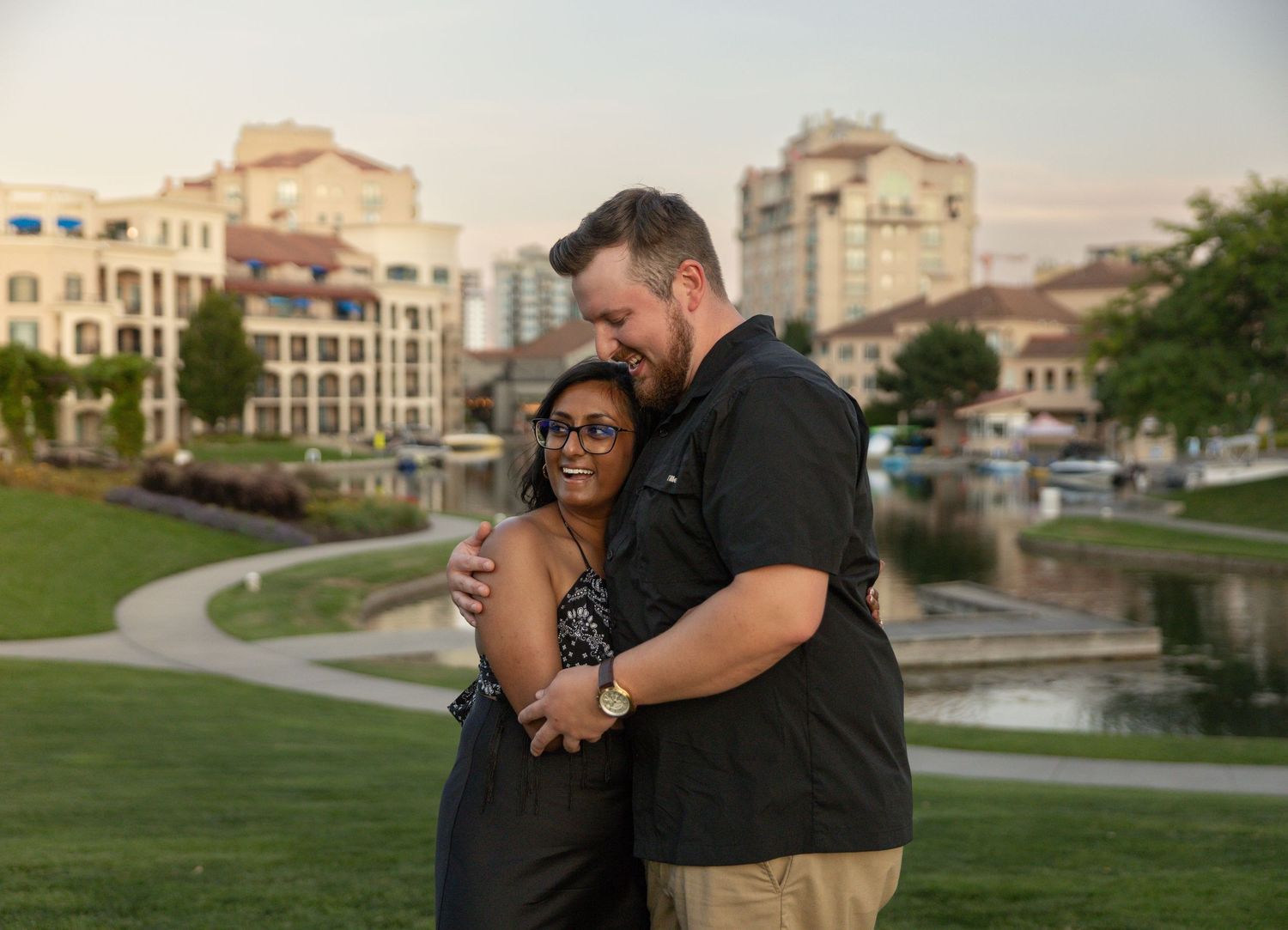Couple embracing and smiling outdoors near a scenic pond with city buildings at sunset in Kelowna, BC