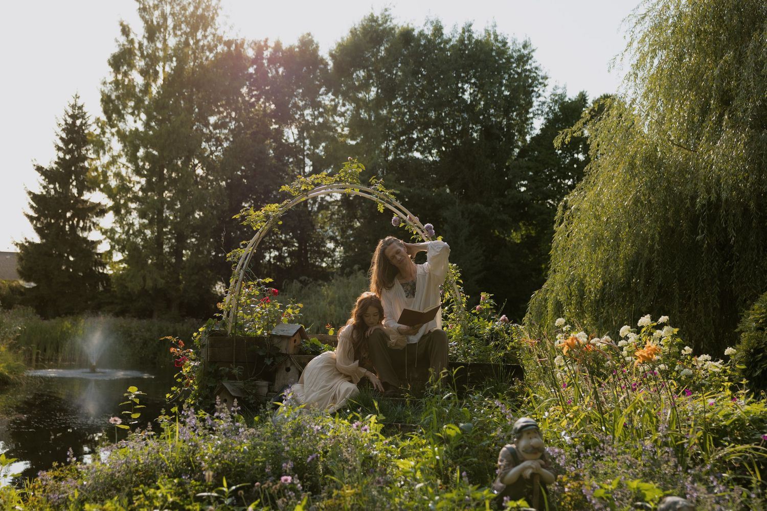 Couple kissing under a floral arch in a lush garden with willow trees and blooming flowers in golden sunlight.