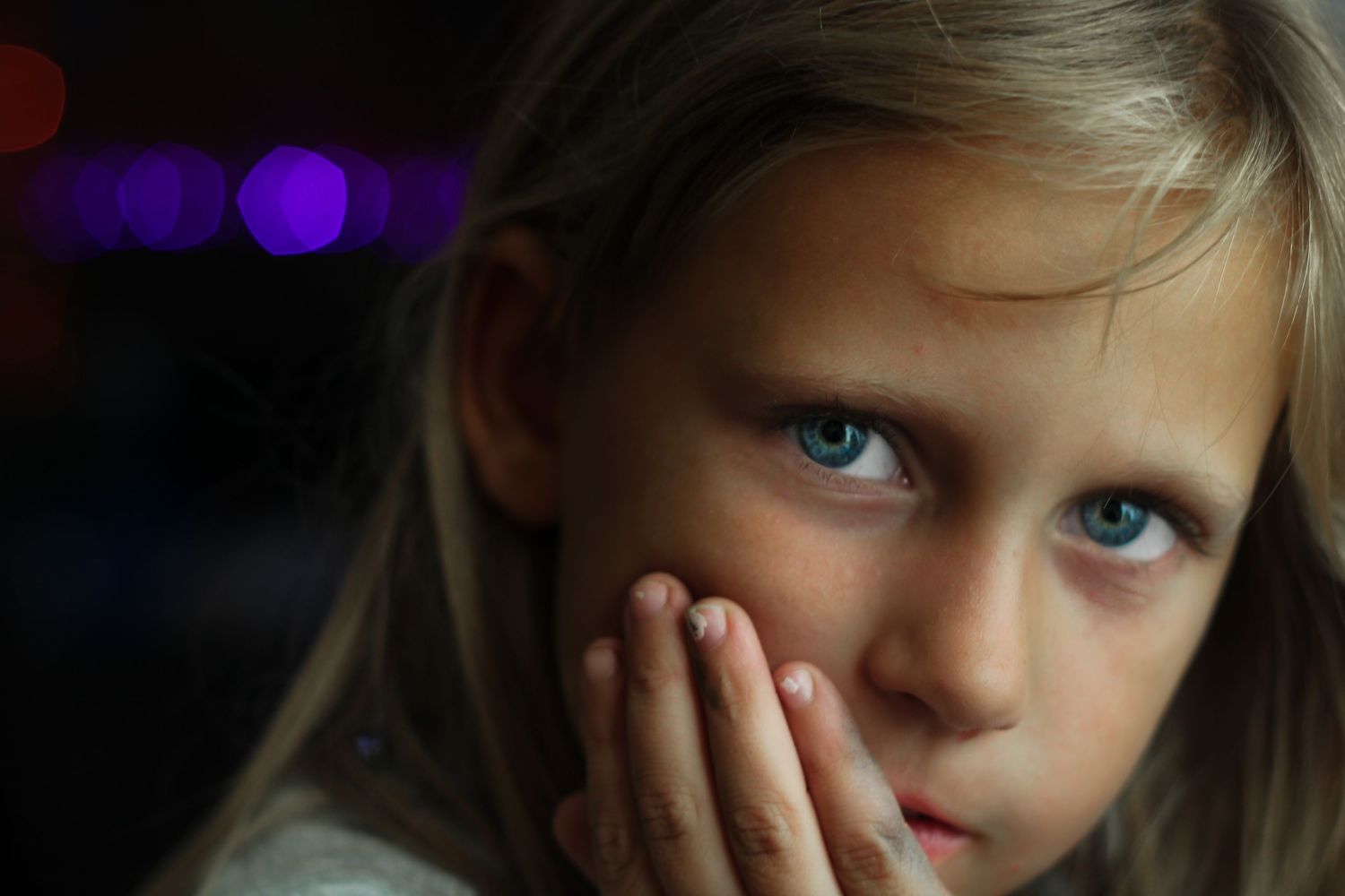 Child with striking blue eyes cupping hands over mouth while looking pensively at camera with blurred lights in background.