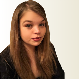 A person with long brown hair wearing a black sweater poses against a light colored wall.