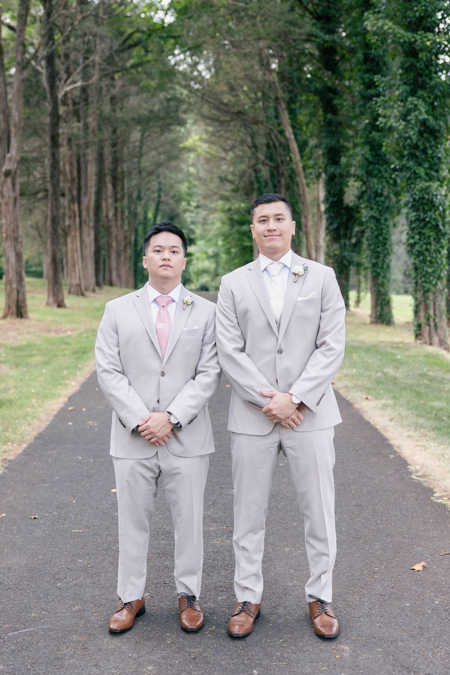 Groomsmen in light gray suits and pink ties pose on tree-lined pathway.Groom and groomsmen wedding outfit inspiration in tan suit.