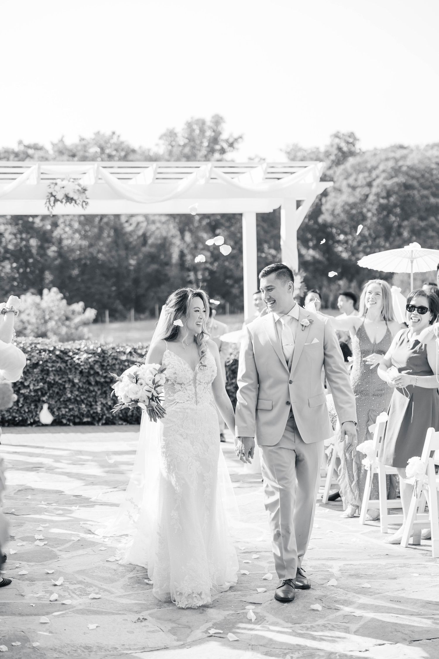 Wedding party walks down an outdoor aisle decorated with white pergola and floral arrangements.