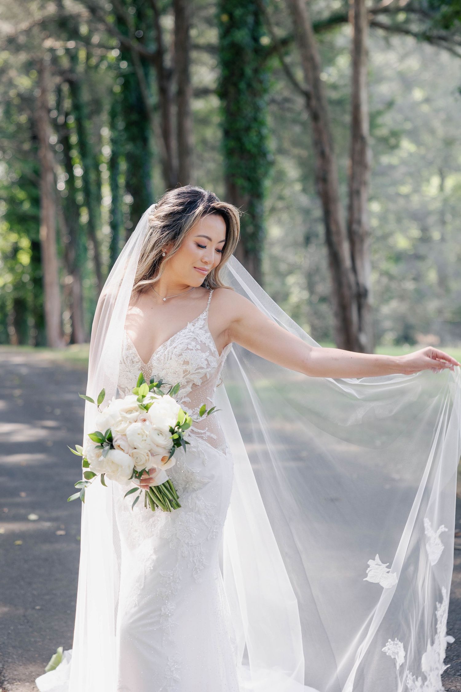 Elegant bride in lace dress holding bouquet among tall trees in a forest setting.