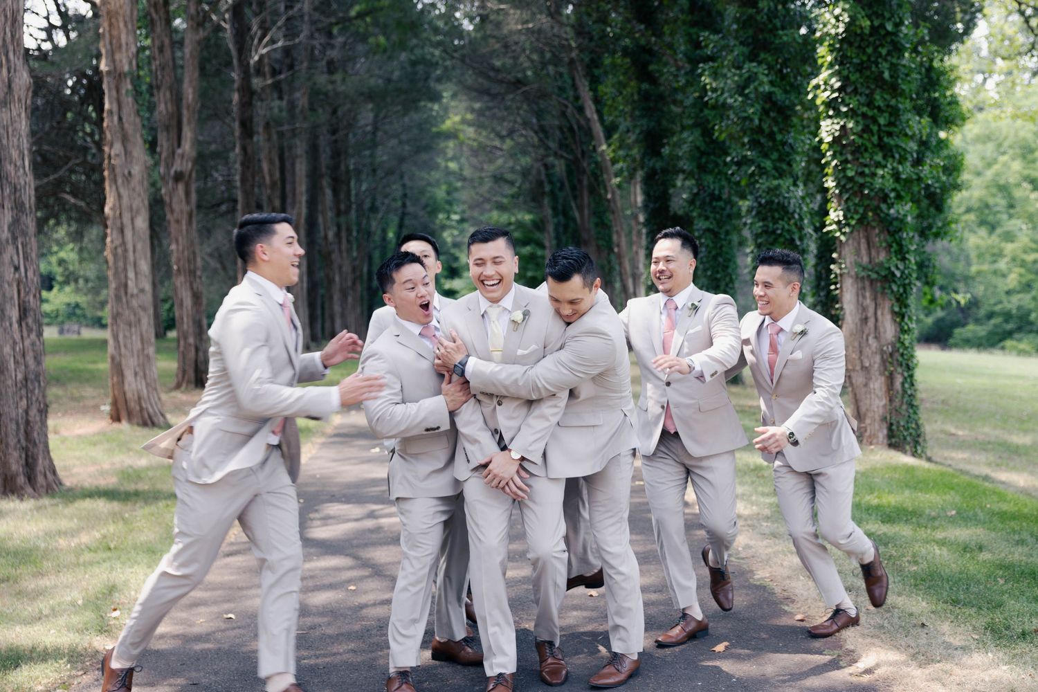 Wedding party members in light suits pose energetically on a forest trail.