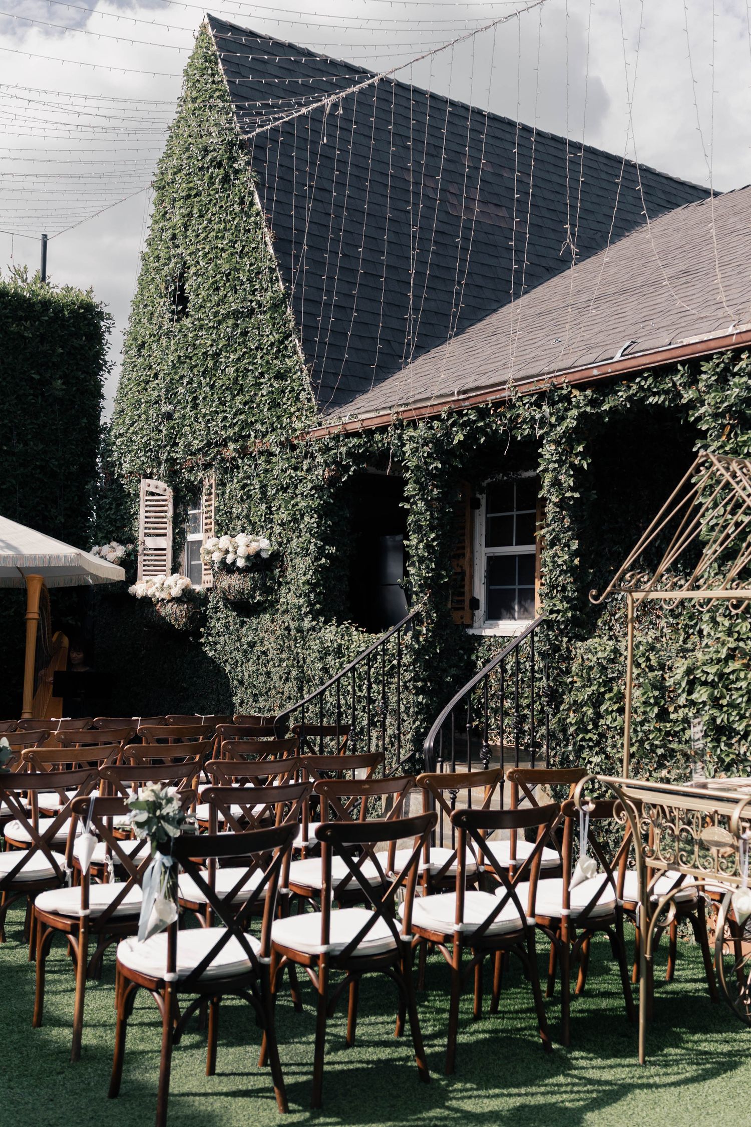 Rows of wooden cross-back chairs set up for outdoor wedding ceremony at ivy-covered venue with rustic charm.
